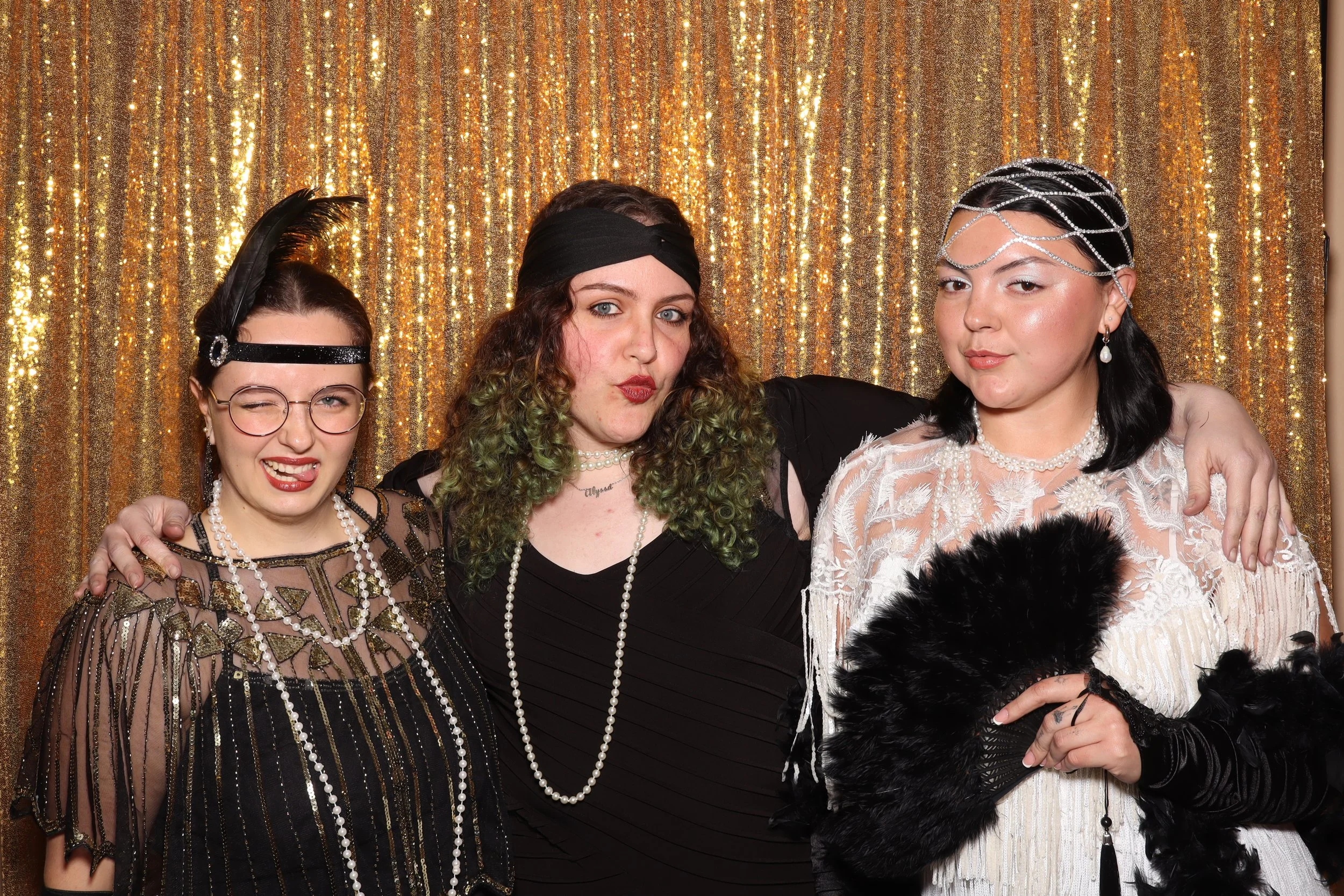 Three women dressed in 1920s-style costumes with pearl necklaces, headbands, and vintage accessories, standing against a gold, glittery backdrop.