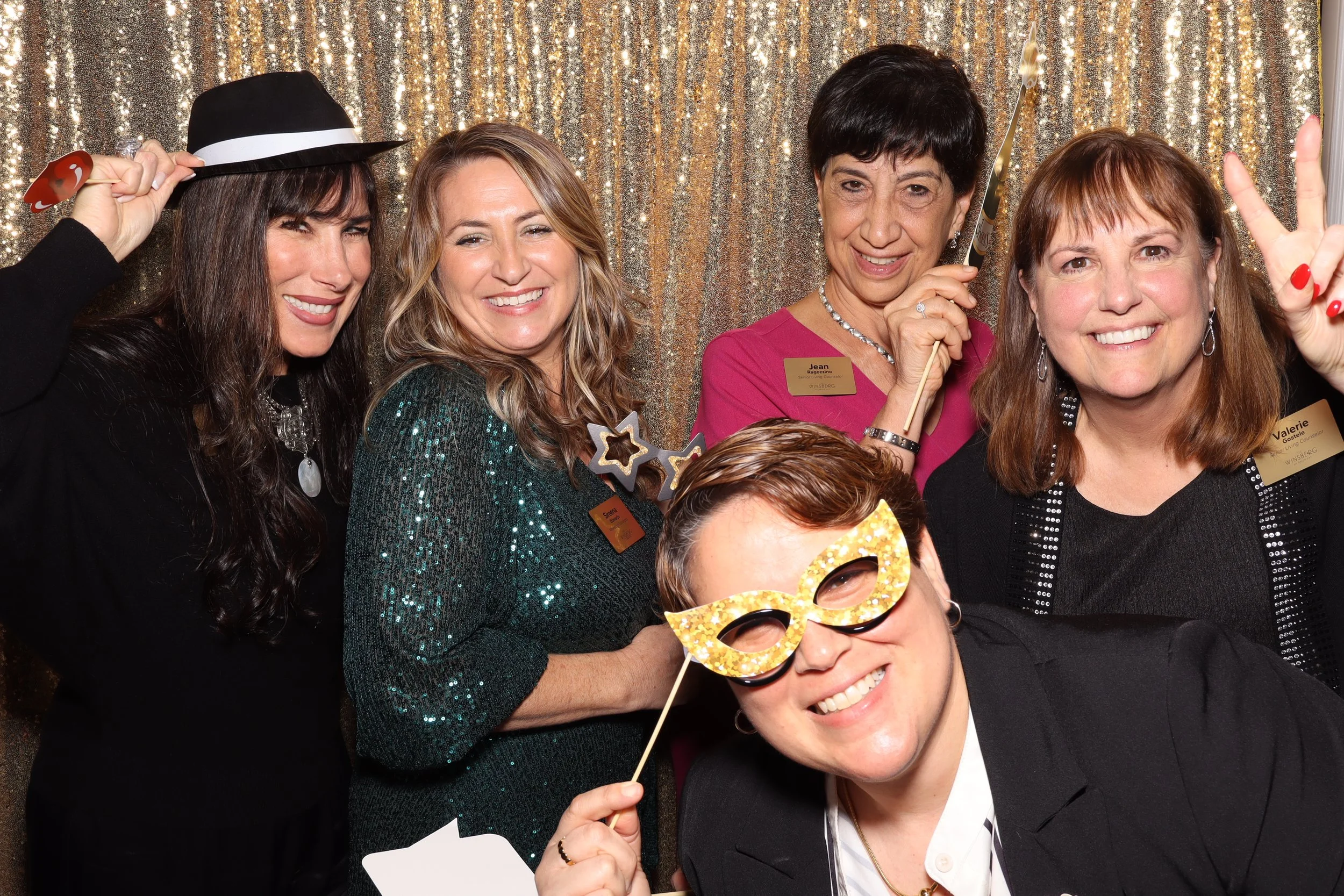 Group of five women celebrating at a festive event, some holding party props like a mask and star-shaped glasses, smiling in front of a glittery gold backdrop.