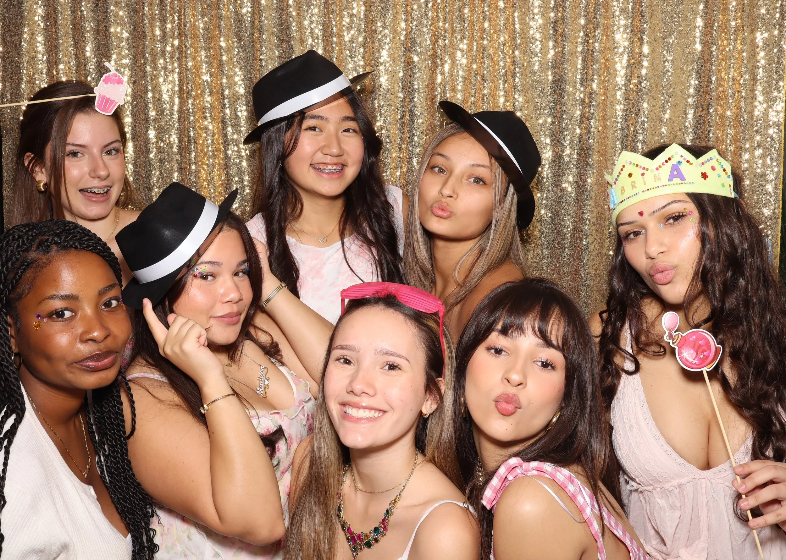 Group of diverse young girls celebrating a birthday, wearing party hats and princess tiaras, posing in front of a gold sequin backdrop, some holding props like a cupcake, a pink perfume bottle, and a sign with the name 'Briana'.