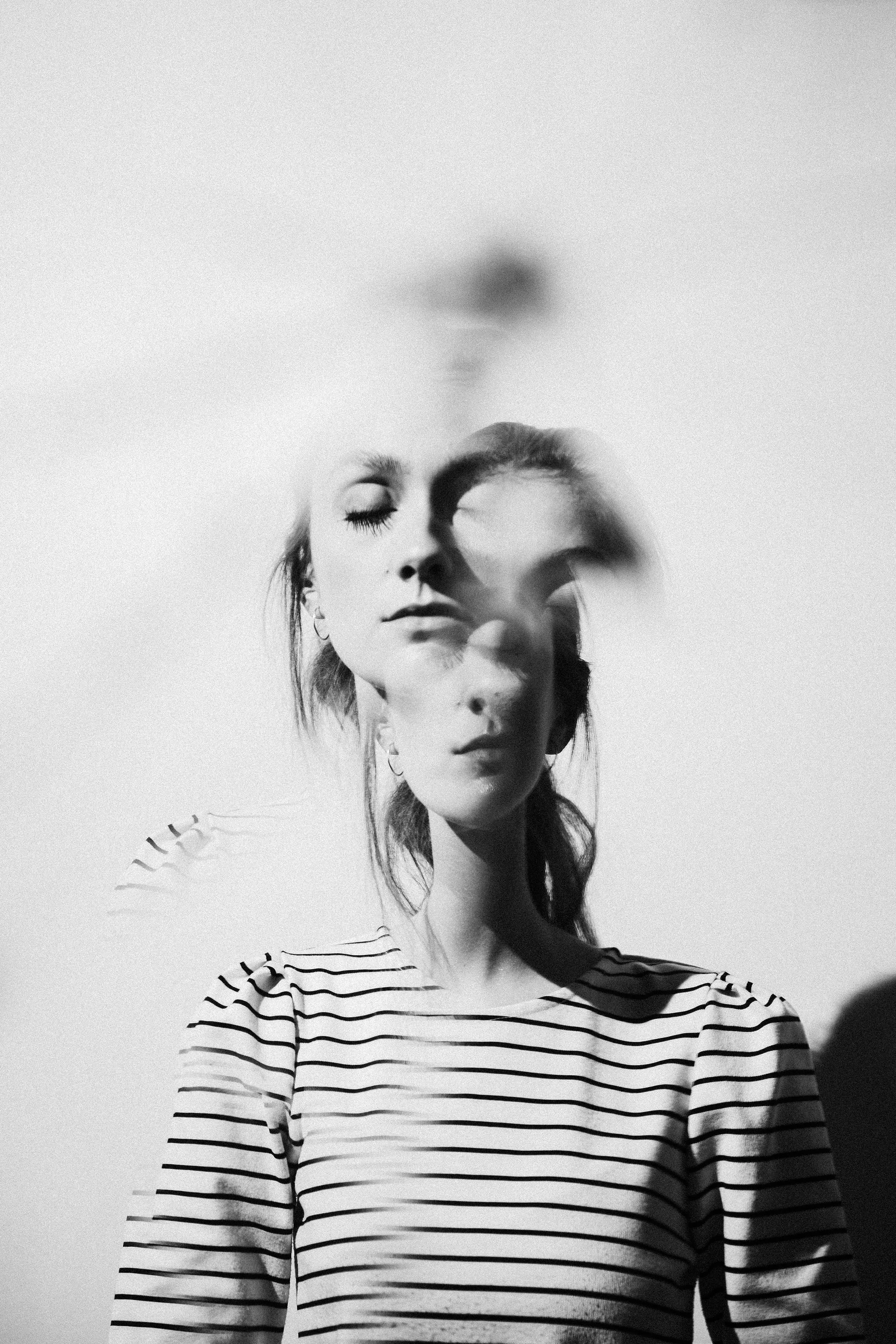 Black and white photo of a woman with her eyes closed, wearing a striped shirt, with a superimposed double exposure effect creating multiple overlapping faces.