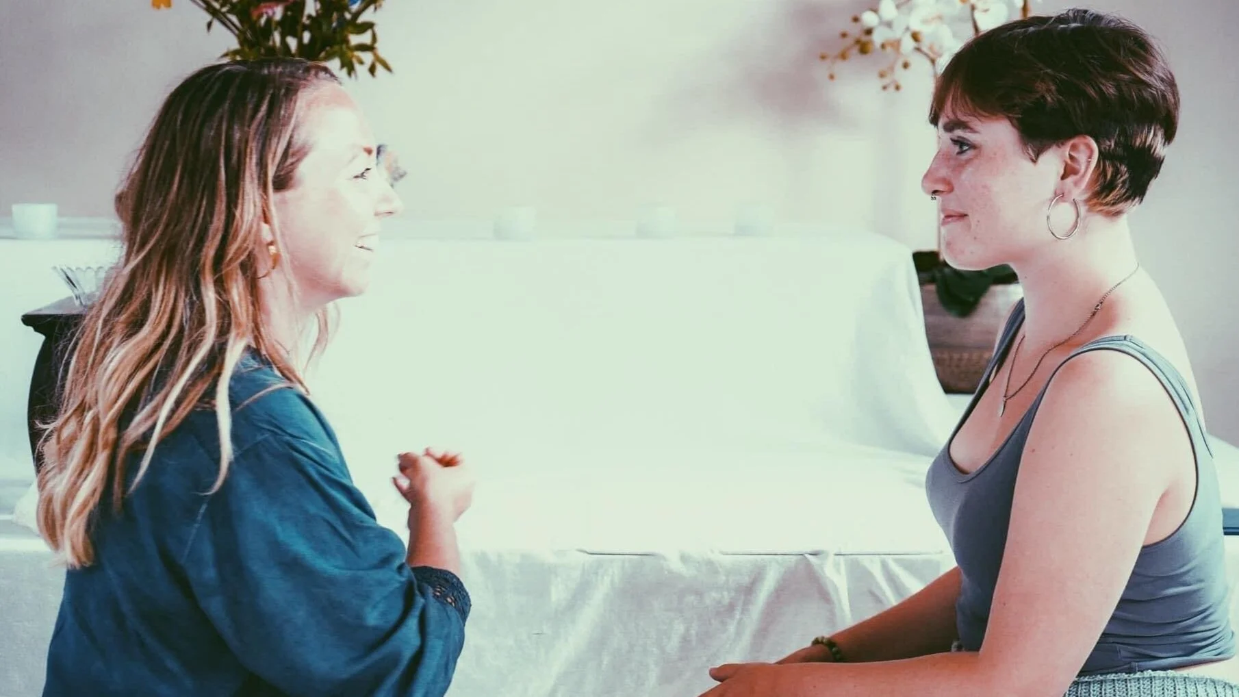 Two women sitting across from each other, engaged in conversation, indoors with a white background and minimal decor.