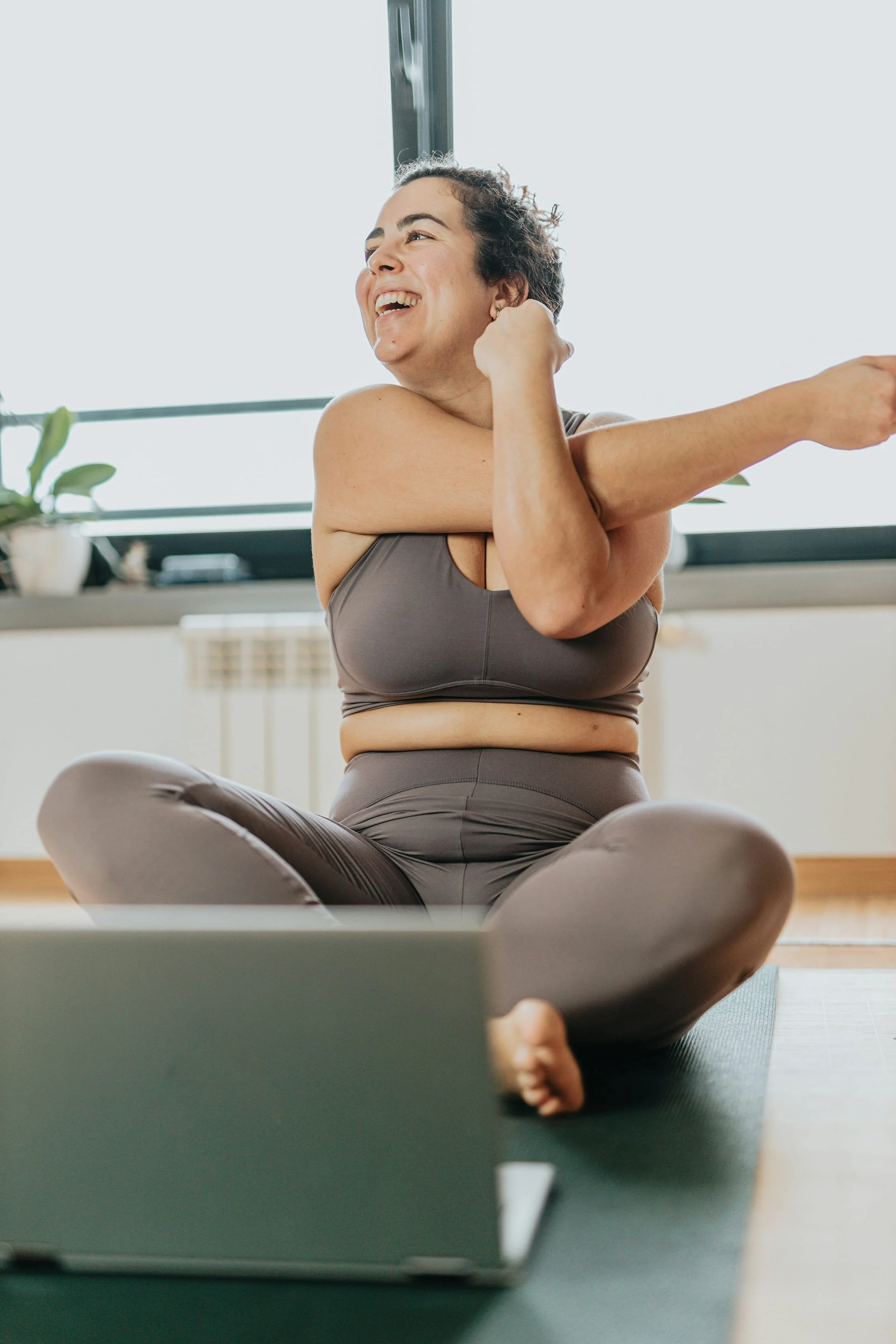 A woman sitting cross-legged on a yoga mat, stretching her arm while smiling during a workout in a bright room with large windows and a plant in the background.