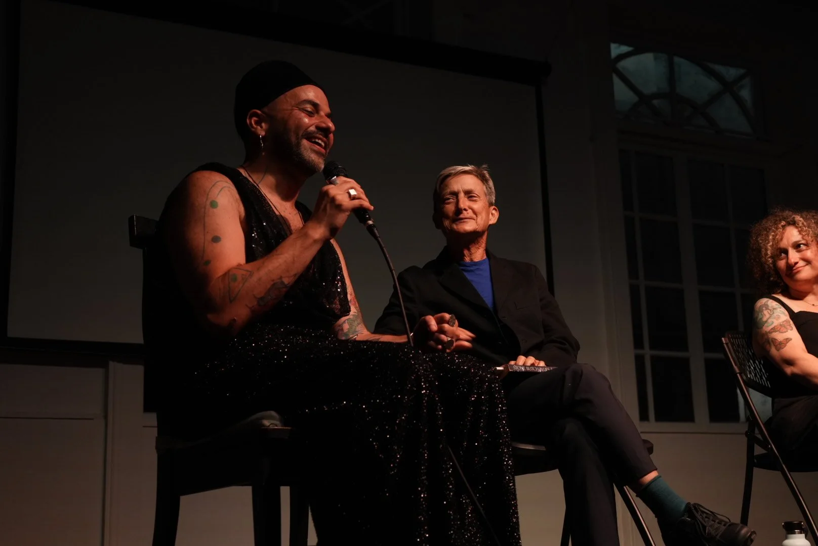 Three people seated on a dimly lit stage during a panel discussion, with a tattooed speaker holding a microphone.