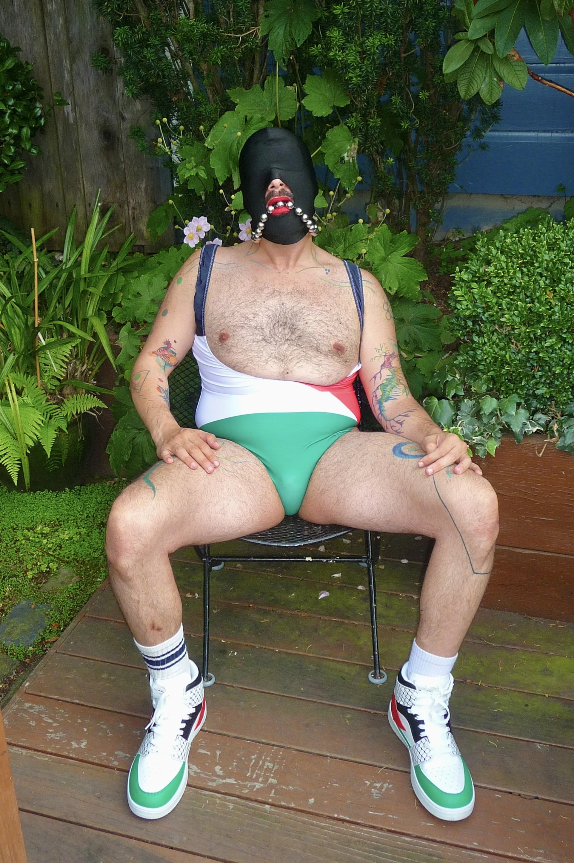 A man sitting outdoors on a wooden deck, wearing a black mask with beads, a swimsuit with the Italian flag colors, white sneakers, and white socks with black stripes, surrounded by green plants.