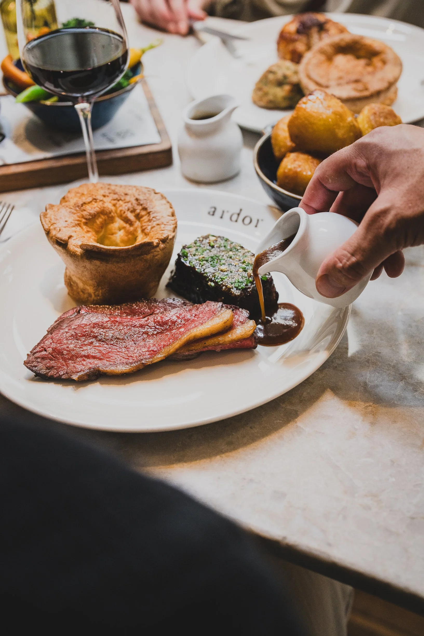A plate of roast beef with Yorkshire pudding and sautéed greens, with gravy being poured over the meat. A glass of red wine is visible in the background. Additional dishes, including potatoes and more Yorkshire pudding, are on the table.