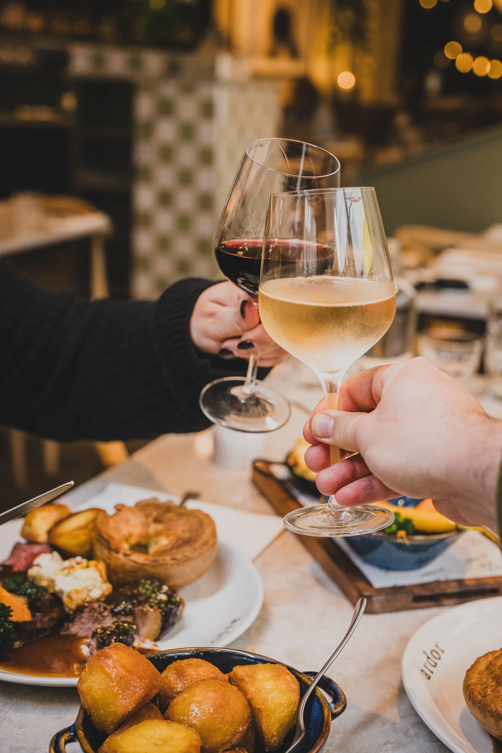 Two people clinking glasses of red and white wine over a table of food at a restaurant.