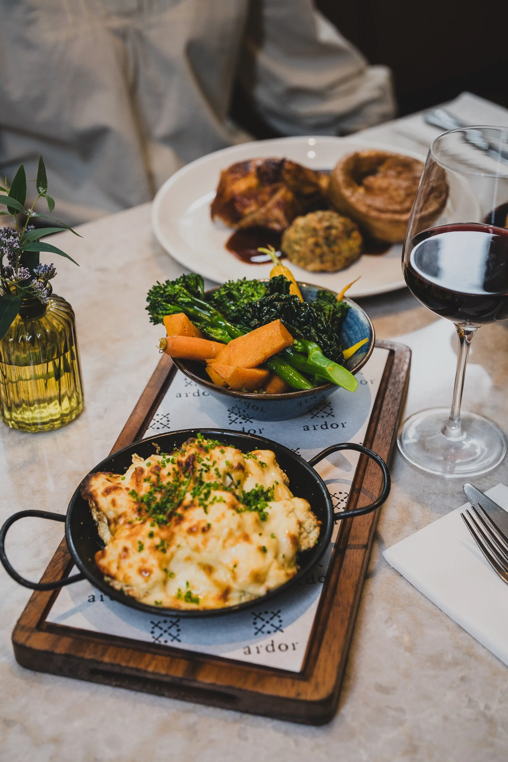 A dining table set with a wooden serving tray holding a black skillet with baked cheesy dish garnished with chives, a small bowl of roasted vegetables including carrots, broccoli, and asparagus, a glass of red wine, and a plate of assorted baked goods in the background.
