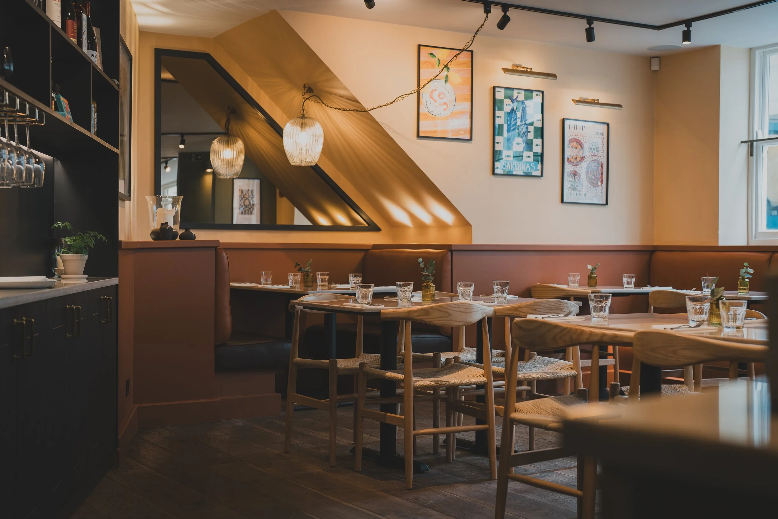 Empty restaurant with tables set with glasses and small vases, framed art on the wall, hanging pendant lights, large window, and a mirror reflecting part of the room.