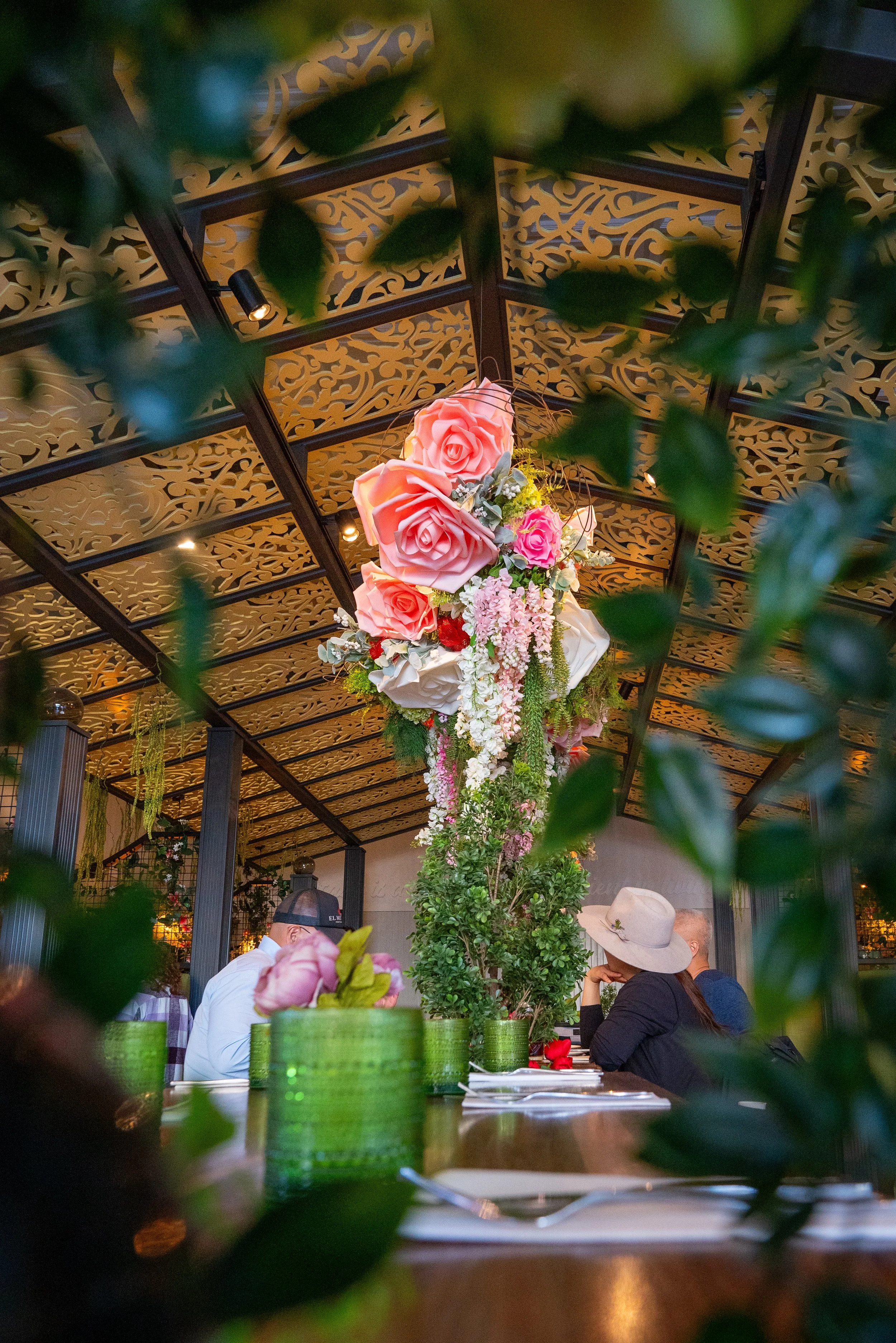 A floral centerpiece with large pink roses and hanging pink and white flowers on a table in the Queenstown Village restaurant in La Jolla, California, seen through green leaves in the foreground.