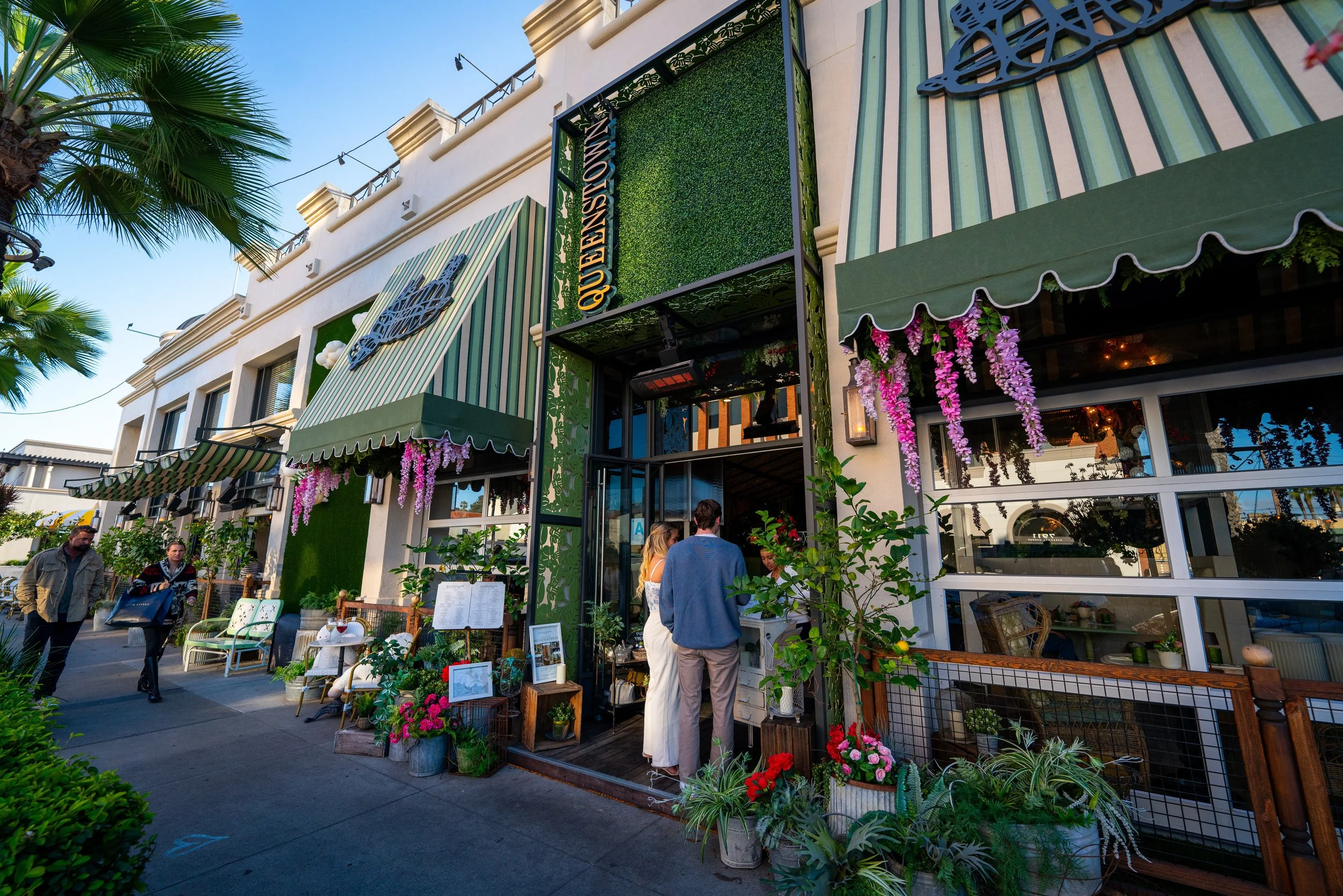People gather outside of Queenstown Village restaurant in La Jolla with green and white striped awnings, lush plants, and pink hanging flowers. There are outdoor seating areas with decorative items and a sign that reads 'QUEENSTOWN'.