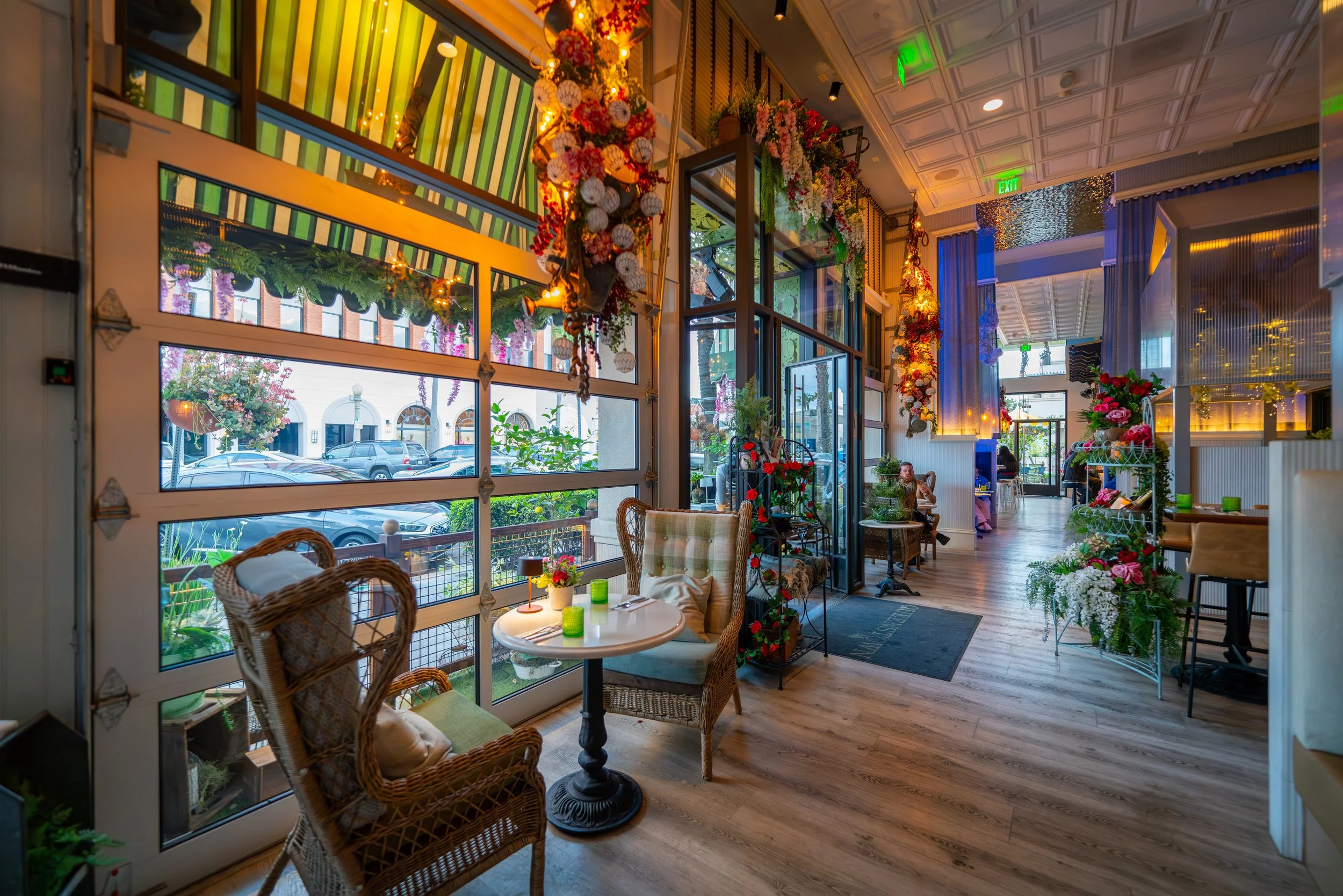 Decorative interior of cozy Queenstown Village restaurant with floral arrangements, colorful lights, wicker chairs, and large windows showing parked cars outside.