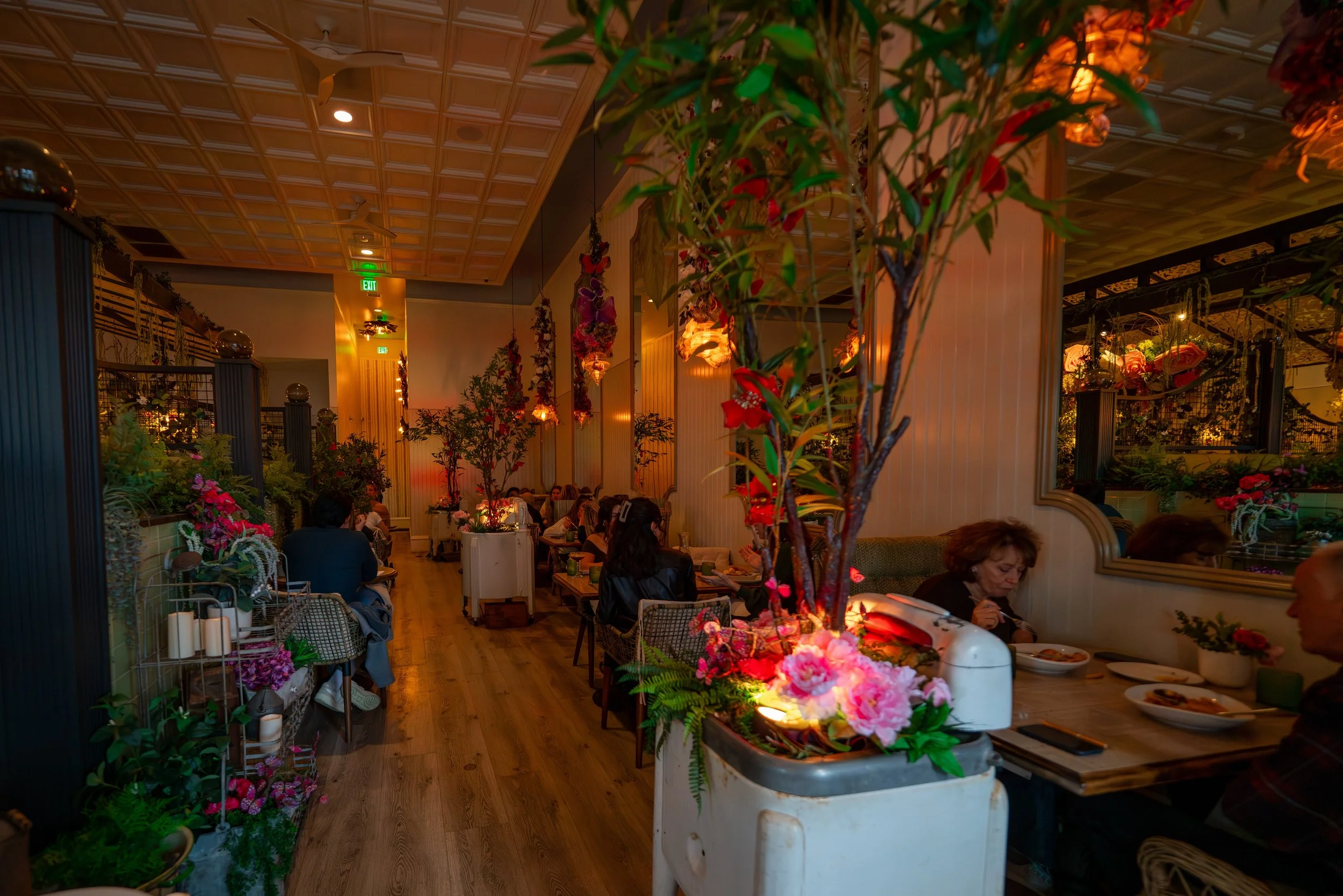 Interior of Queenstown Village restaurant with floral and plant decorations, warm lighting, and several groups of people dining.