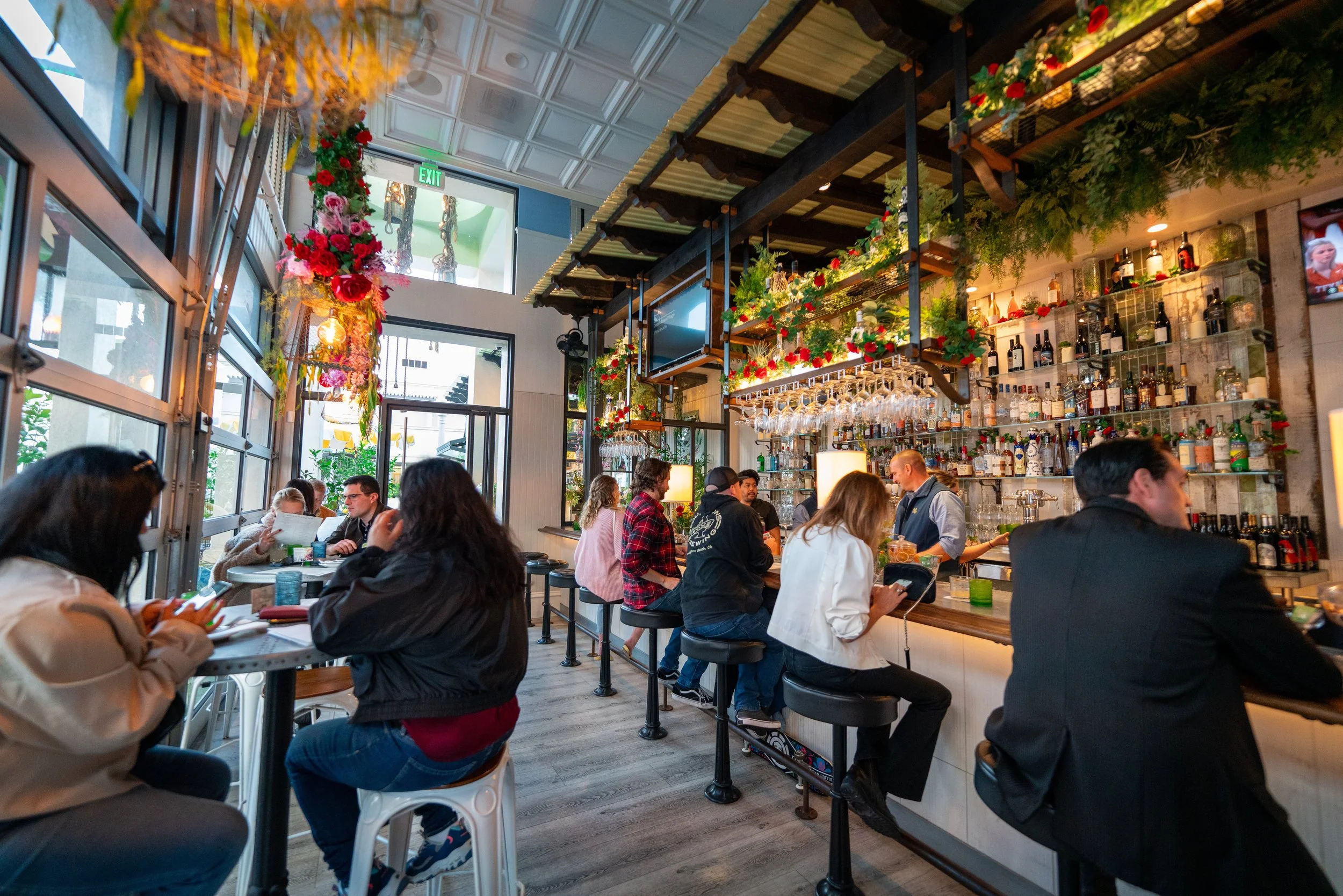 People sitting at tables and a bar inside a restaurant decorated with flowers and greenery, with large windows and a staircase in the background.