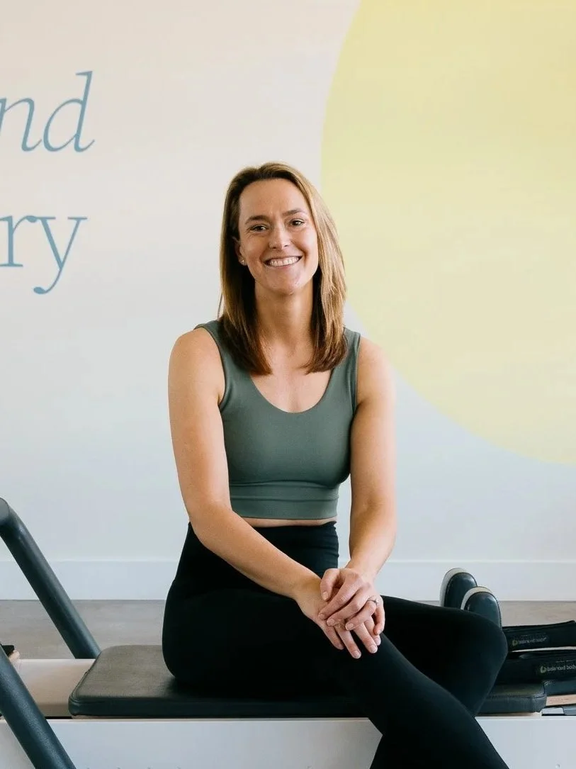 A woman with shoulder-length brown hair smiling, sitting on a Pilates reformer machine in a fitness studio.
