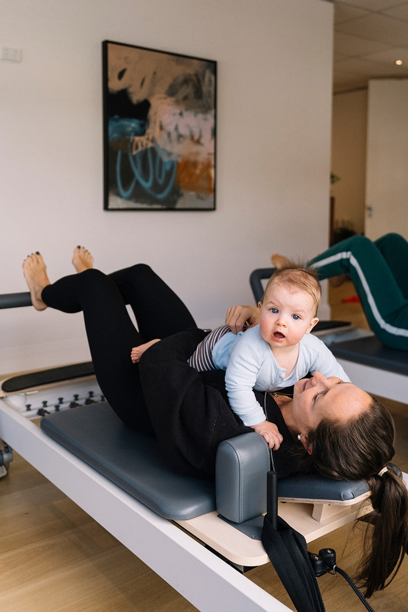 Mother laying down on pilates reformer with baby on her belly.