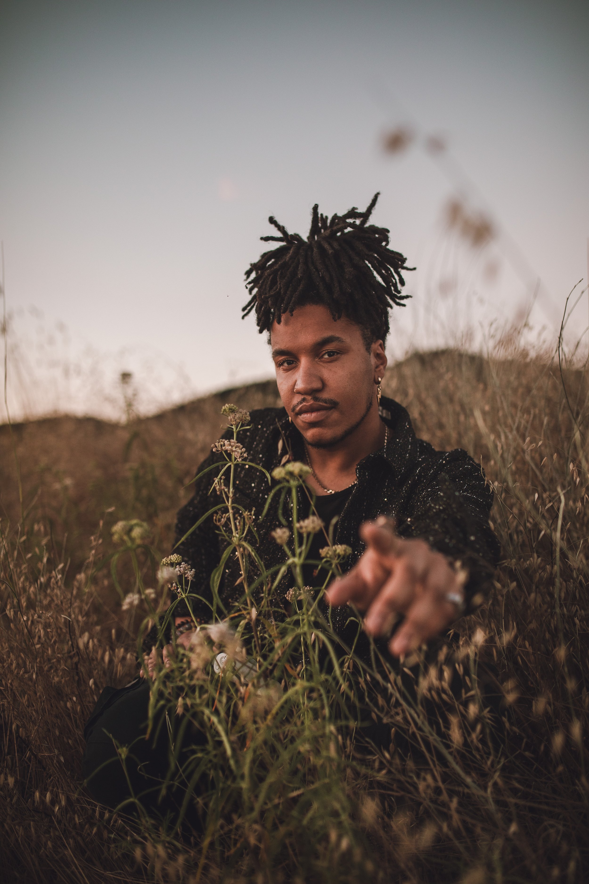 A portrait of a man with dreadlocks sitting in a field, pointing towards the camera, tall grass and plants around them, wearing a dark outfit.