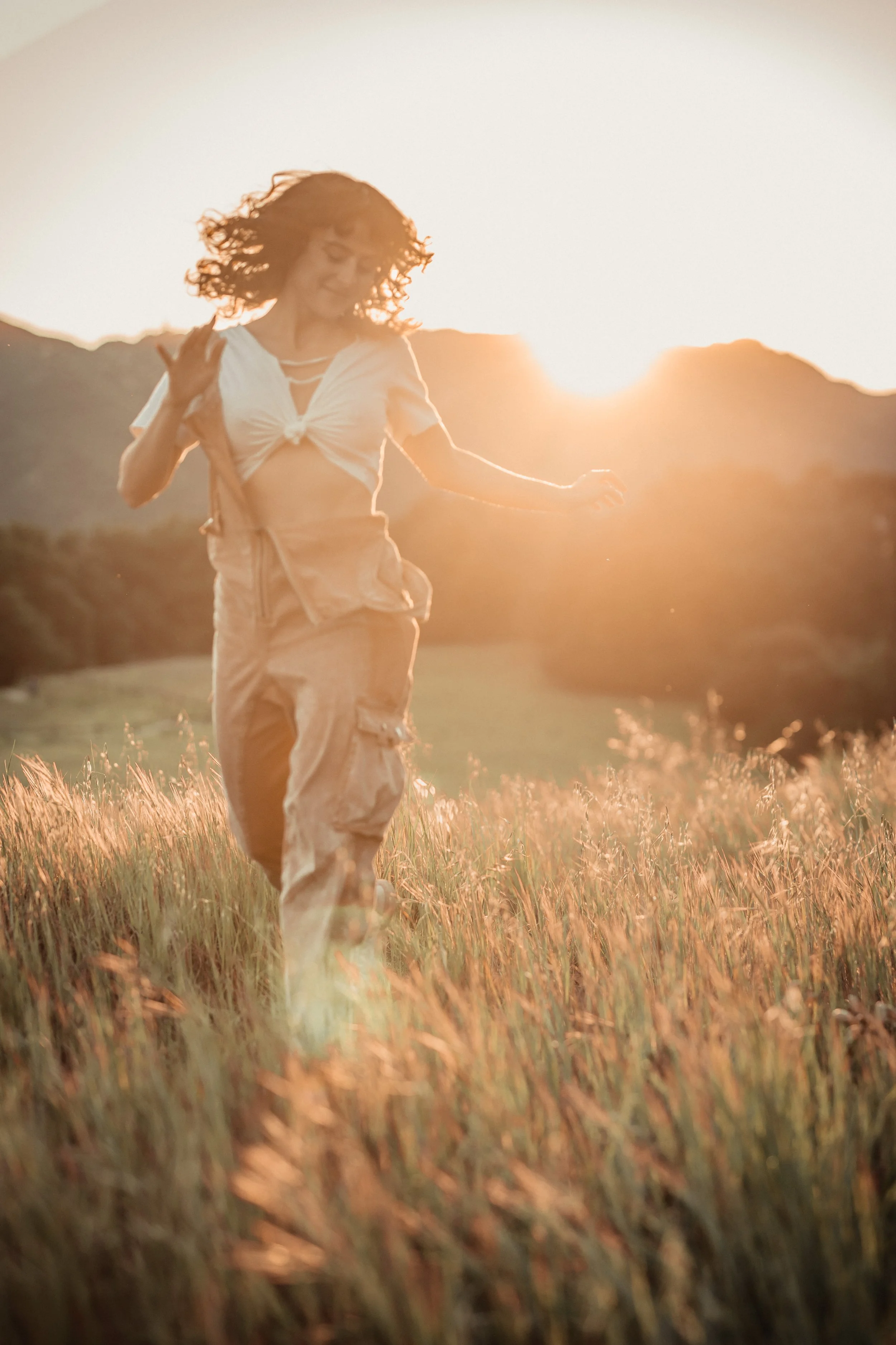 A portrait of a young brunette woman in overalls, enjoying a sunny day in a grassy field with the sun setting behind mountains.