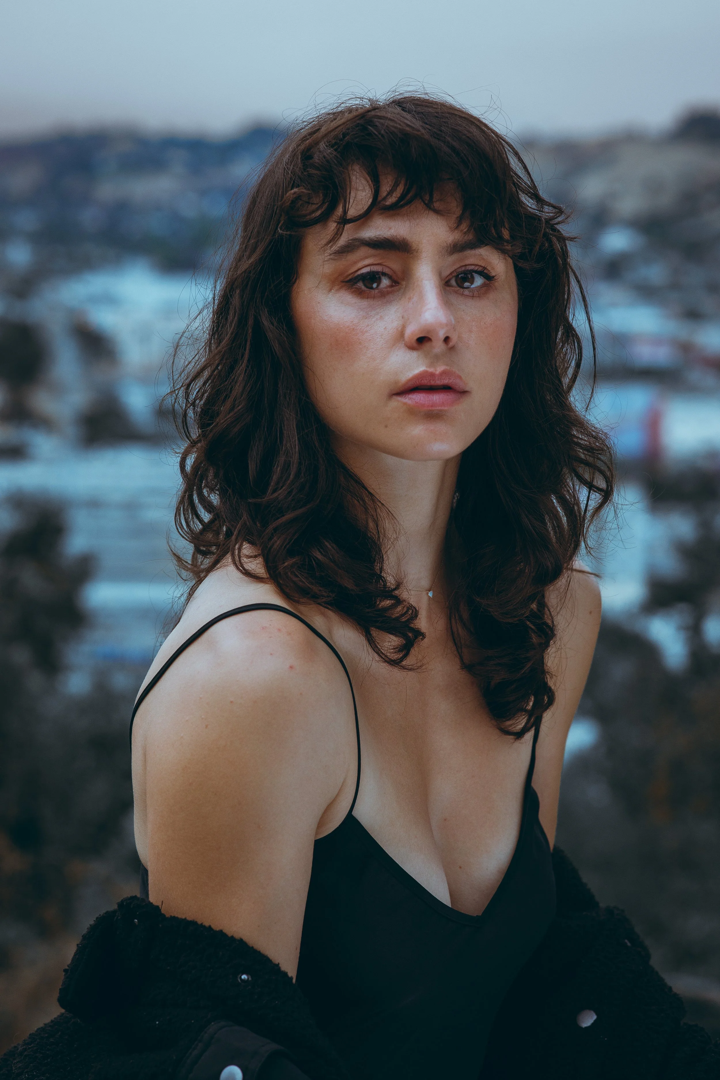 A woman with dark curly hair wearing a black spaghetti strap top and a black jacket, standing outdoors with a blurred natural background.