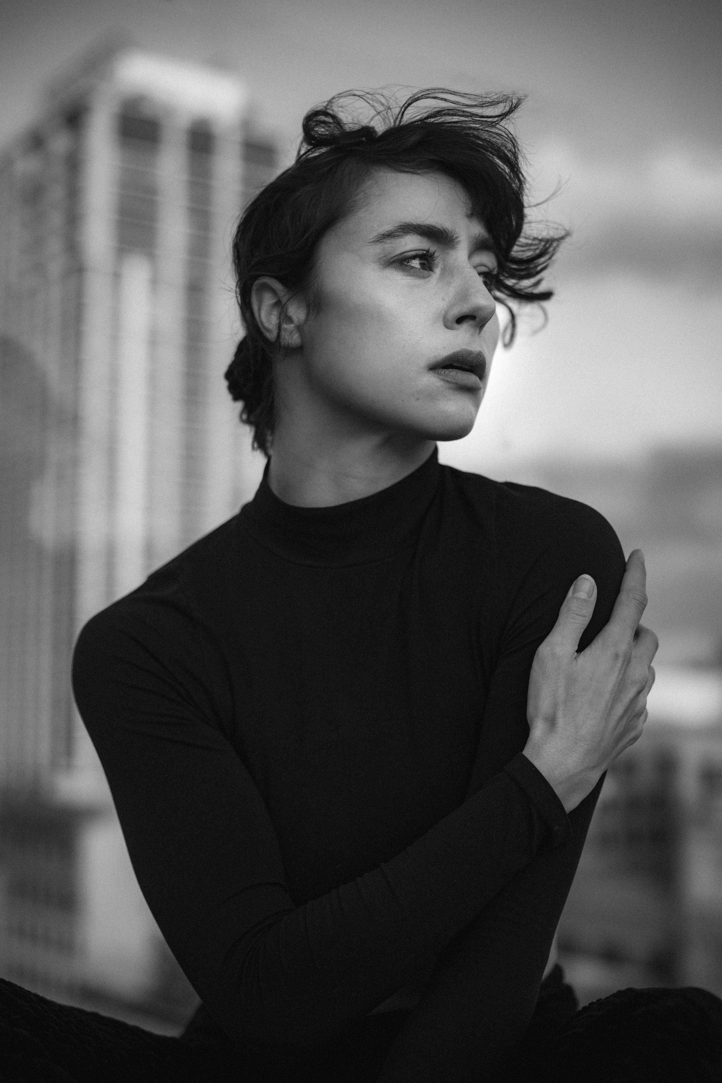 A black and white portrait of a young woman in a black turtleneck with wind-tousled hair, against a blurred urban background, looking to the side with a thoughtful expression.