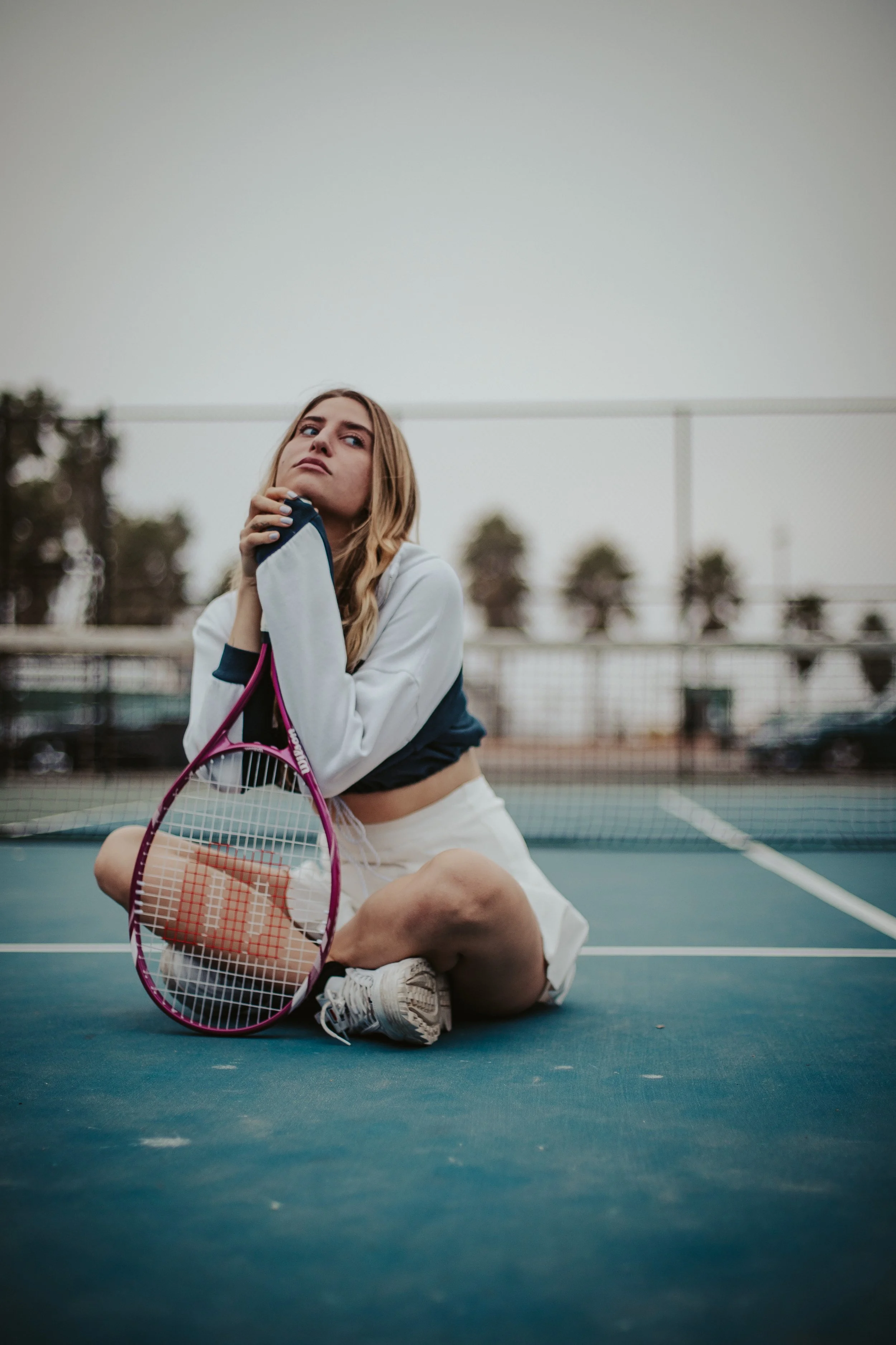 A portrait of a young woman sitting on tennis court holding a tennis racket, looking upward.