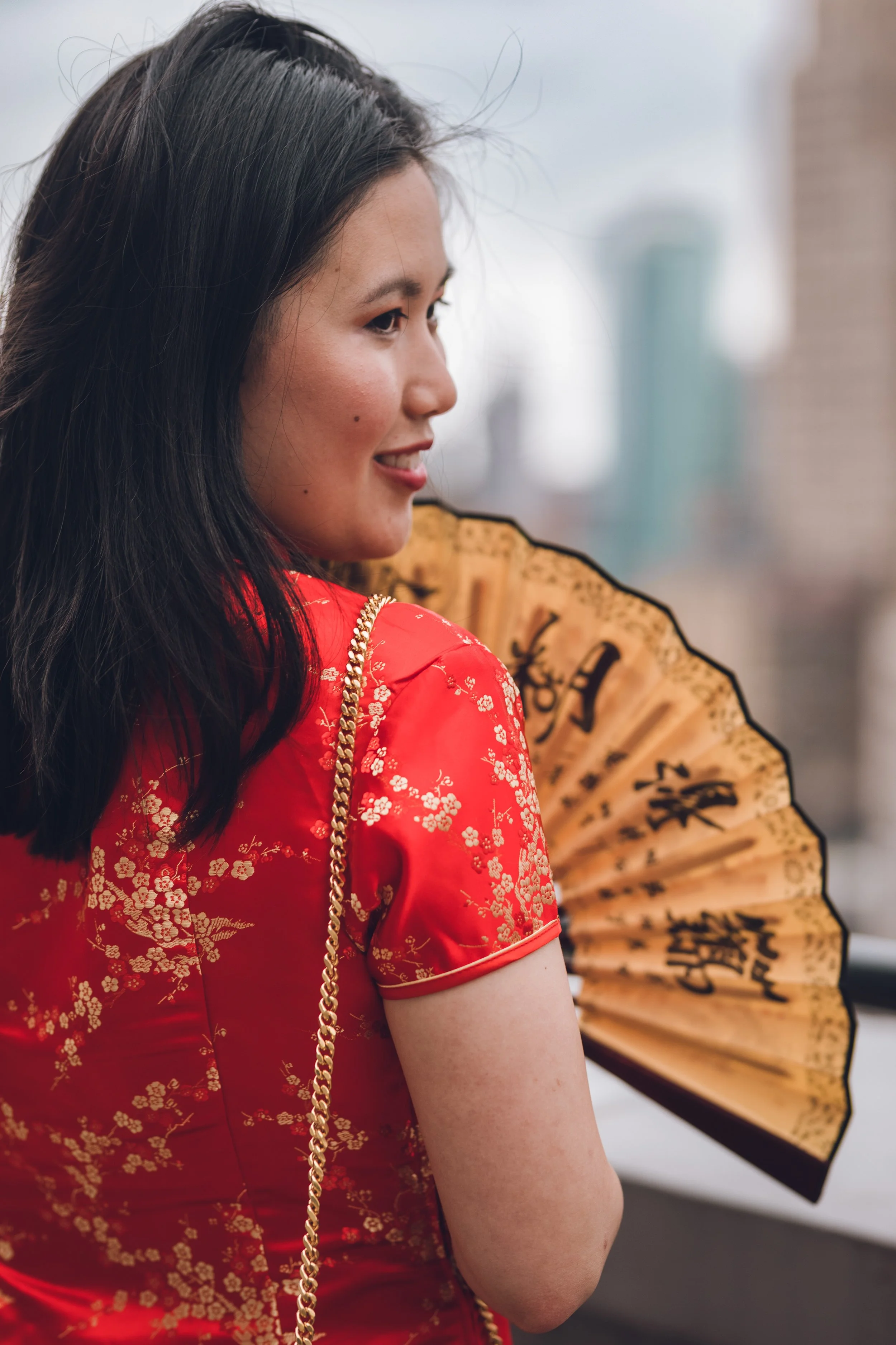 A portrait of a young woman with dark straight hair in red dress holding a decorative fan with Chinese characters, in front of a cityscape background.