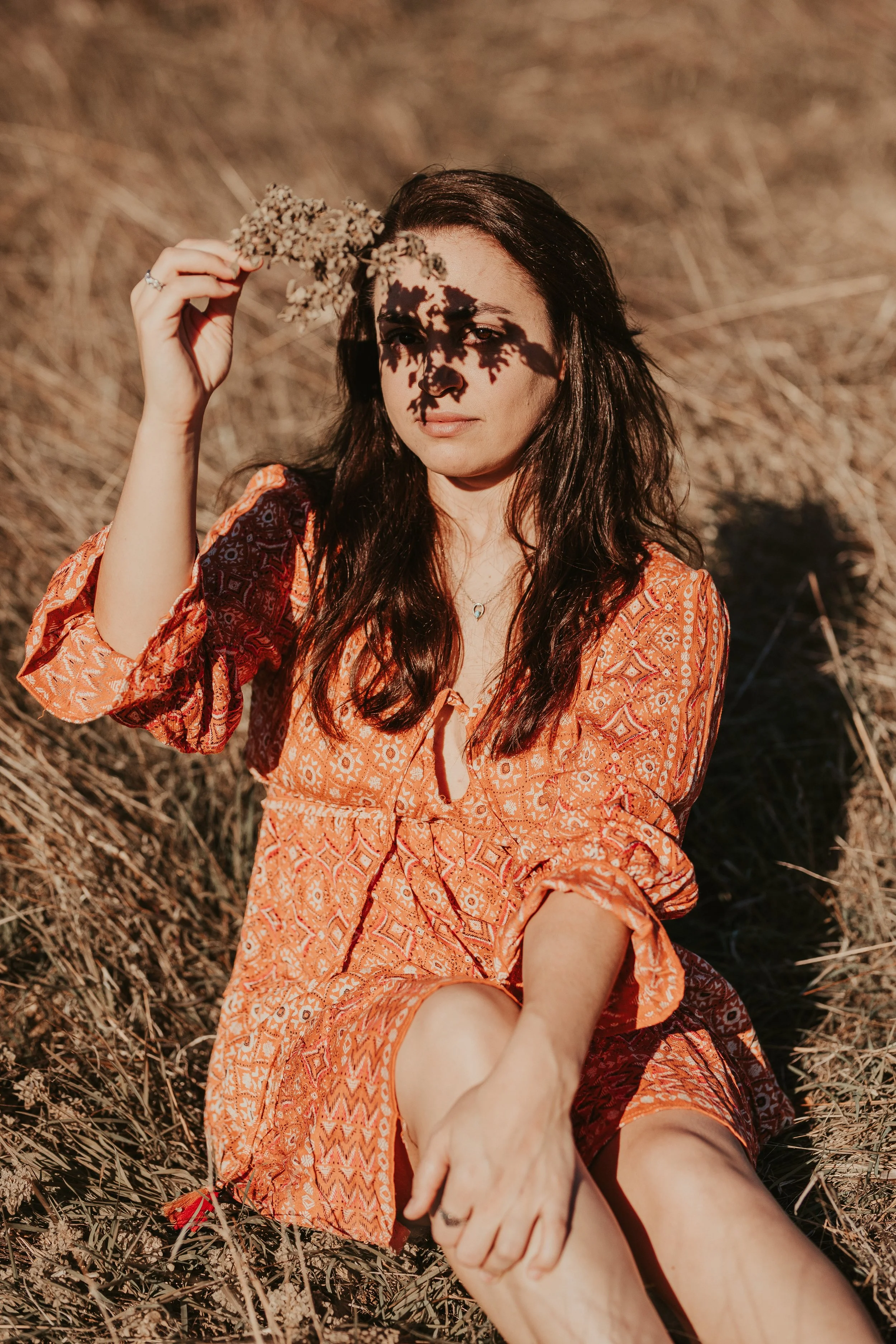A portrait of a young woman in orange patterned dress holding dried flowers over face, casting shadow, sitting in dry grass.
