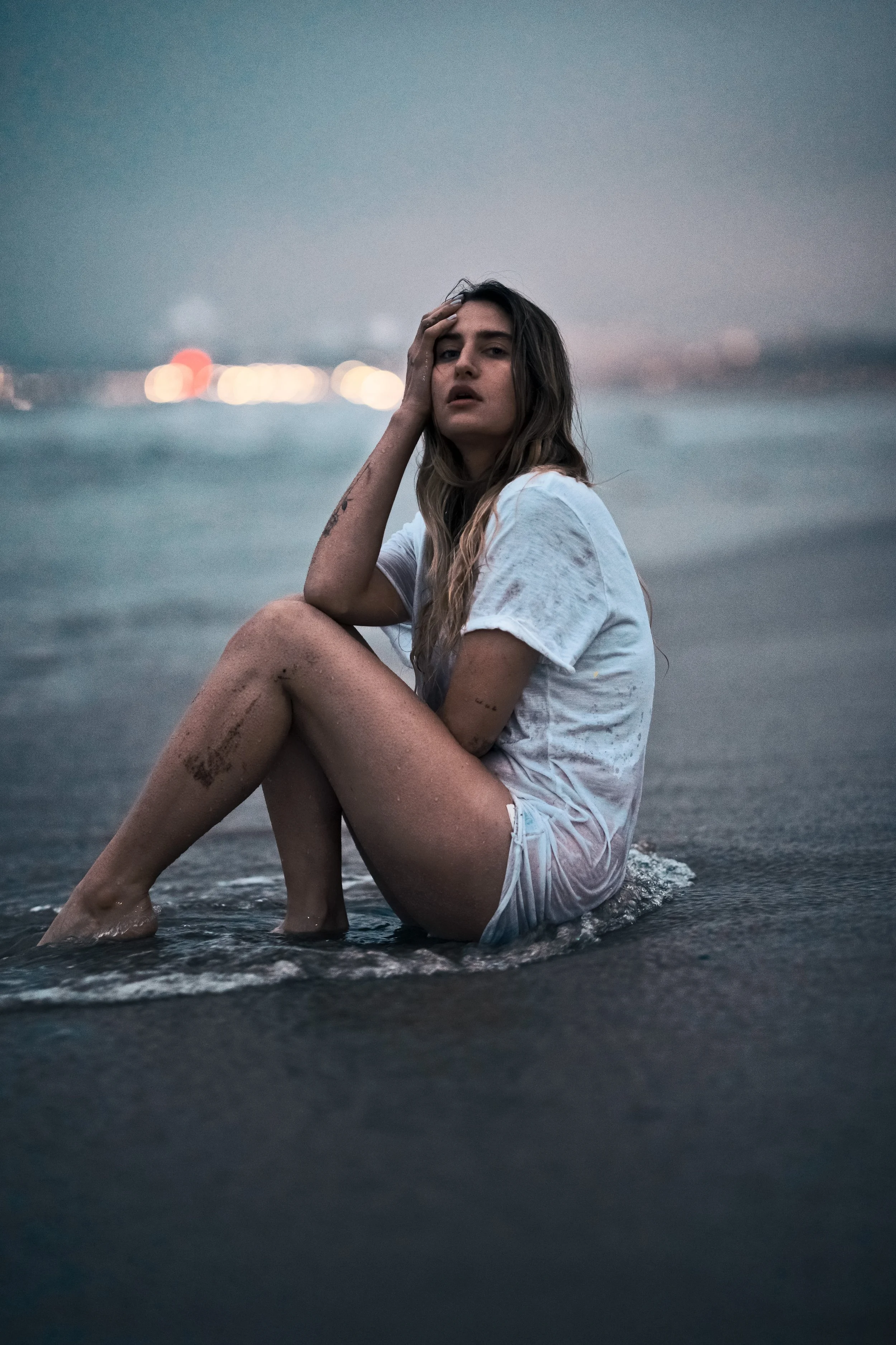 A portrait of a young woman sitting on a beach in a white t-shirt, with wet hair, during dusk.