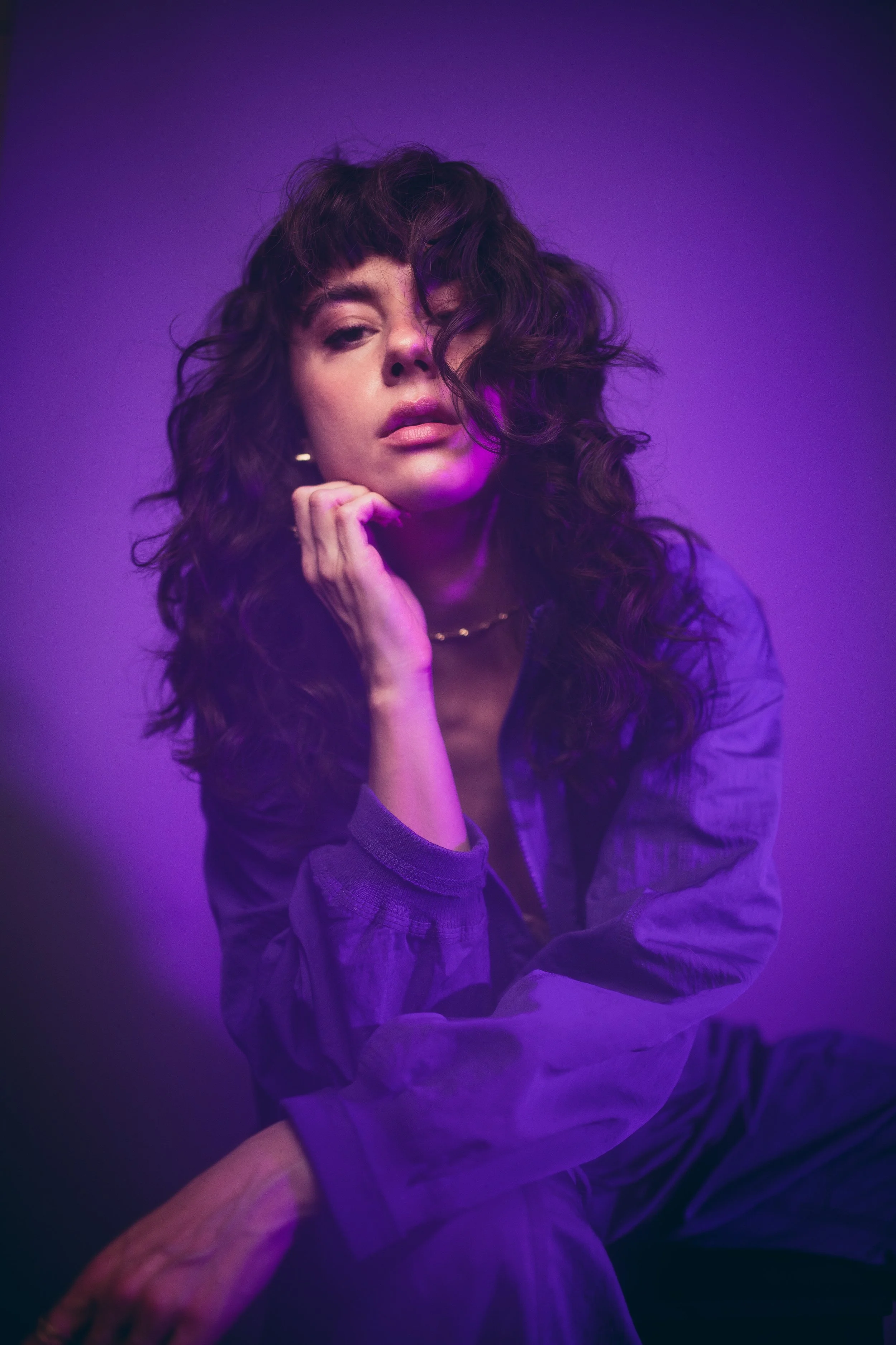 A studio portrait of a young woman with dark, curly hair posing under purple lighting, wearing a purple jumpsuit.