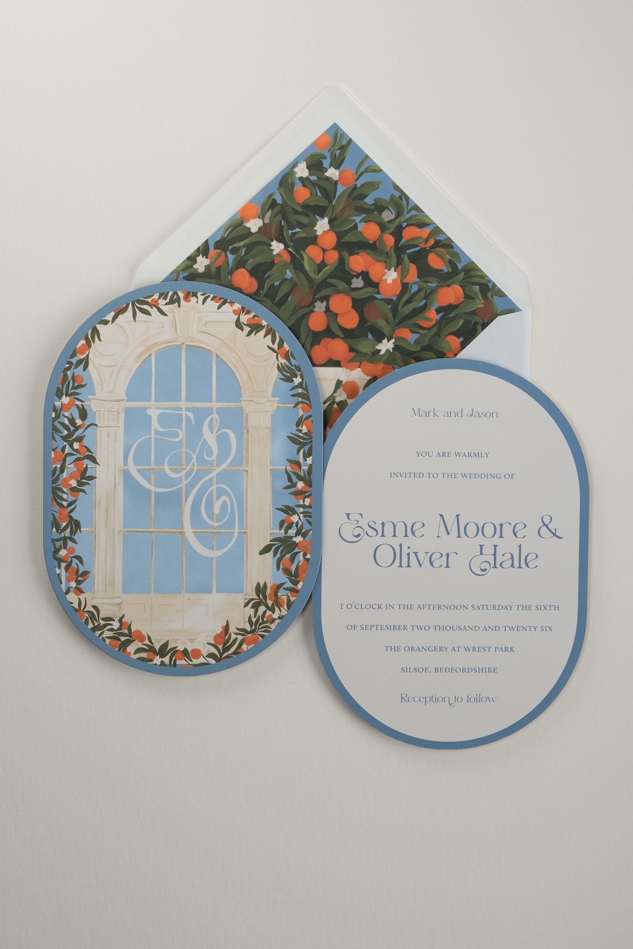 Front and reverse view of the double-arched Orangery wedding invitation laid over a white envelope with an illustrated liner featuring lush orange bush, green leaves, and citrus fruits.