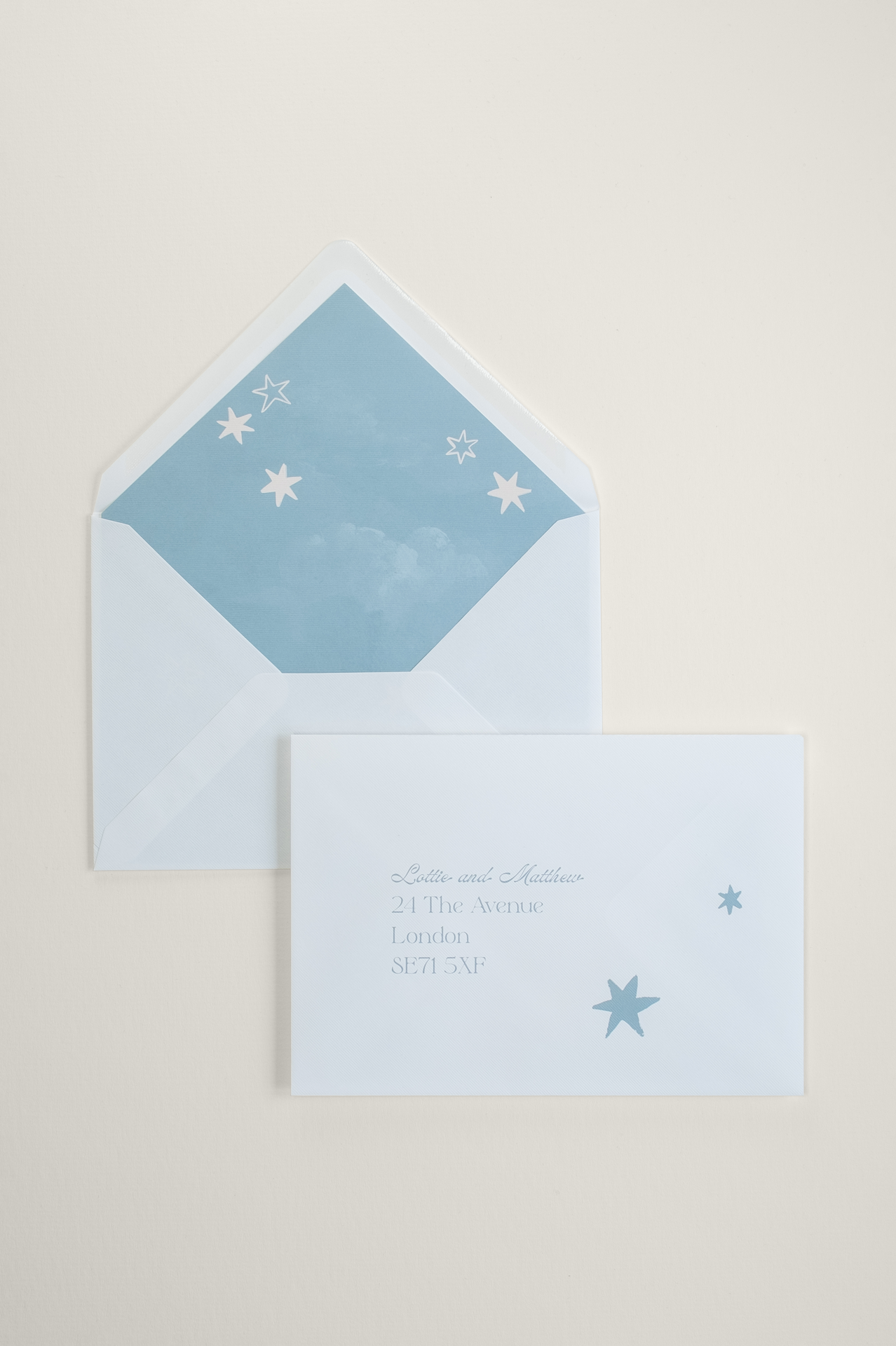 Custom printed wedding envelope front with soft blue guest addressing and star illustrations, layered over an open envelope revealing a matching blue sky liner.