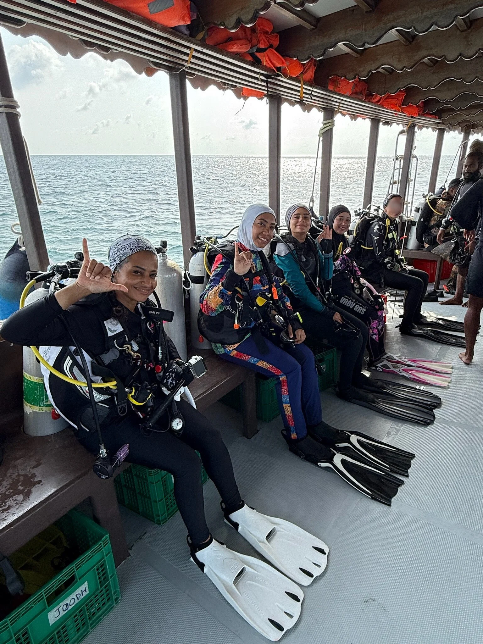 Group of five women in scuba gear sitting on a boat near the ocean, smiling and making hand signs, with scuba tanks, fins, and wetsuits.
