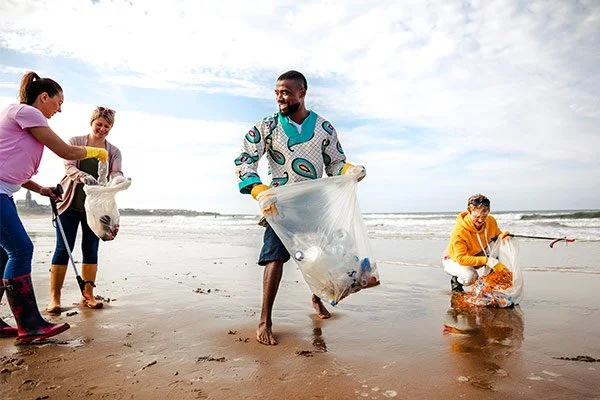 Group of five people cleaning up trash on the beach, some holding trash bags and picking up litter from the sand.