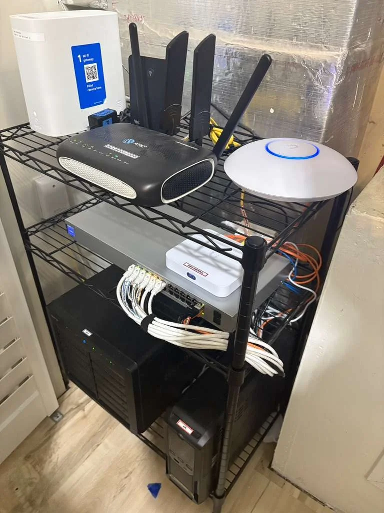 Networking equipment on a black metal shelf, including routers, switches, a Wi-Fi gateway, a wireless access point, and power supplies.