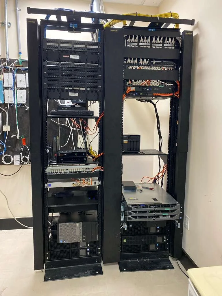 Two tall server racks filled with computer hardware and networking equipment, including switches, servers, and power supplies, with cables running between components.