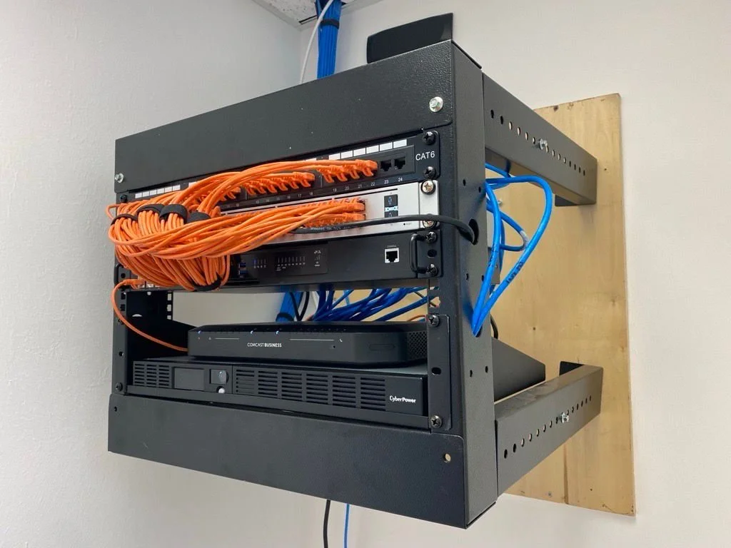 Network equipment rack with orange and blue Ethernet cables connected to switches and a CyberPower uninterruptible power supply, mounted on a wall in a server room.