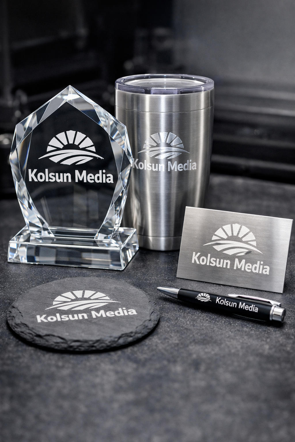 Various branded office items on a dark surface, including a crystal award, travel mug, business card, coaster, and pen, all with the 'Kolsun Media' logo and branding.