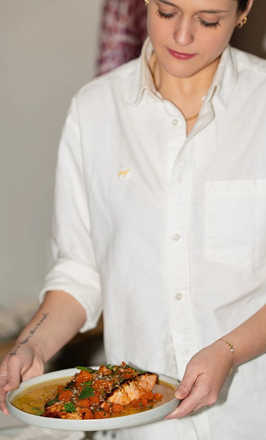 Person holding a plate with a piece of salmon topped with a vegetable mixture, garnished with herbs.