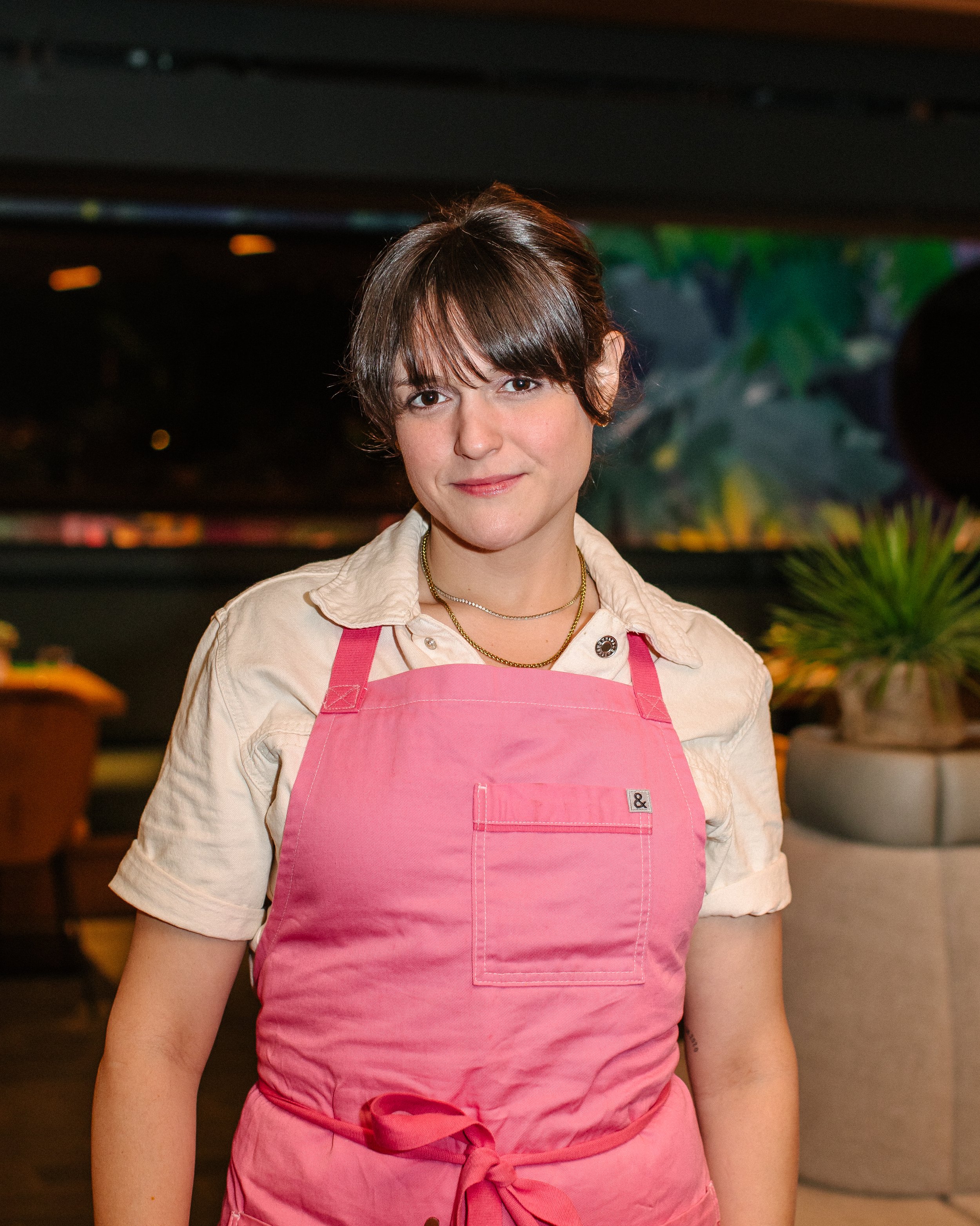 A woman with short brown hair wearing a pink apron over a beige shirt, standing in an indoor setting with plants and blurred background.