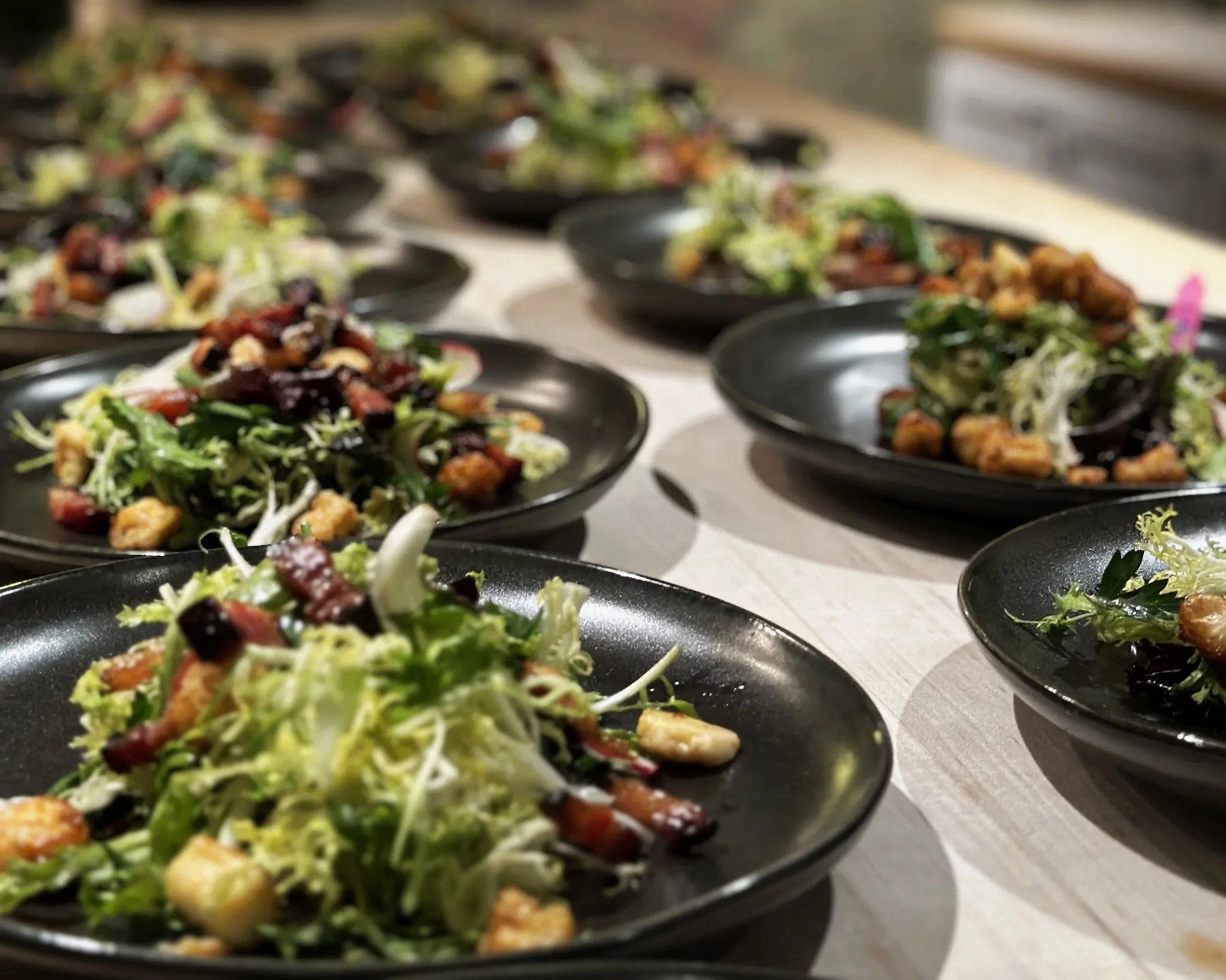 Multiple black plates arranged in rows, each with a fresh mixed salad, on a white surface.