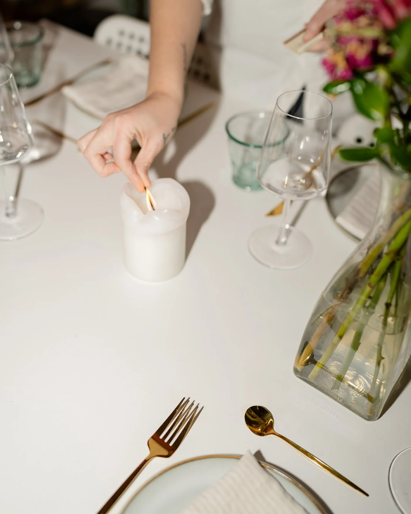 A person lighting a candle on a white dining table decorated with flowers, wine glasses, a gold spoon and fork, and a vase with flowers.