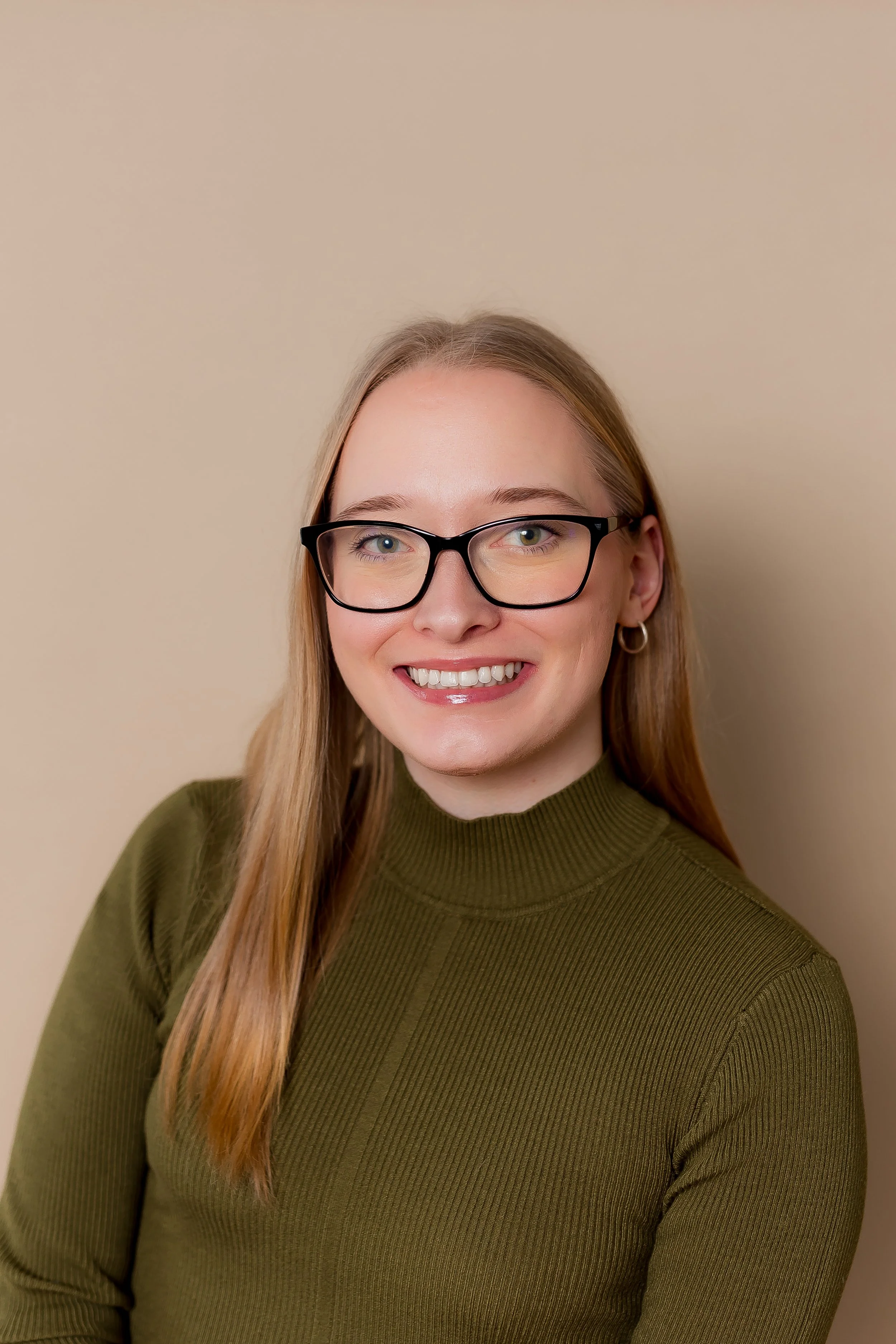 A young woman with long blonde hair wearing glasses and a green ribbed turtleneck sweater, smiling against a beige background.