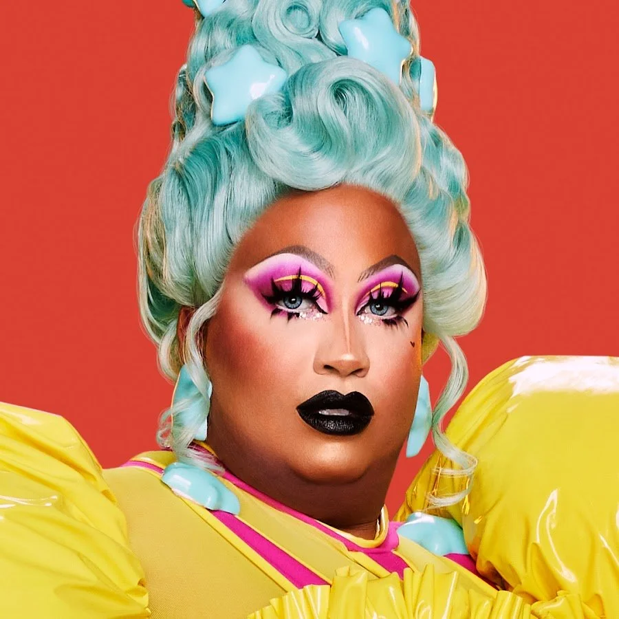 A person with vibrant makeup and teal hair styled in an elaborate updo, adorned with light blue star-shaped decorations. They are wearing bold pink and purple eyeshadow, dramatic eyeliner, black lipstick, and a bright yellow outfit with puffy sleeves. The background is a solid orange.