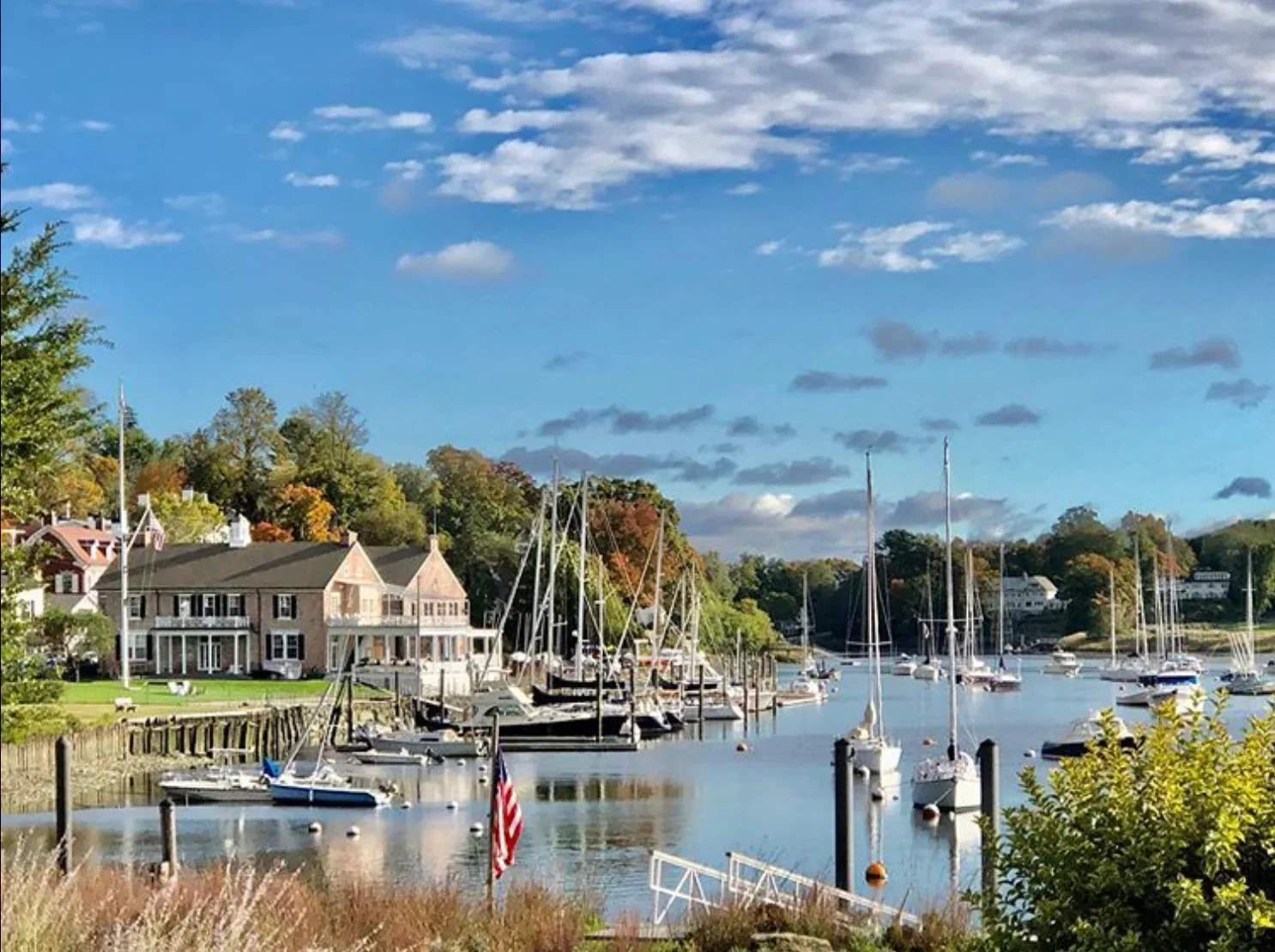Scenic waterfront view of a peaceful marina with sailboats docked along calm water, traditional New England-style homes in the background, and trees with early autumn colors under a bright blue sky with scattered clouds