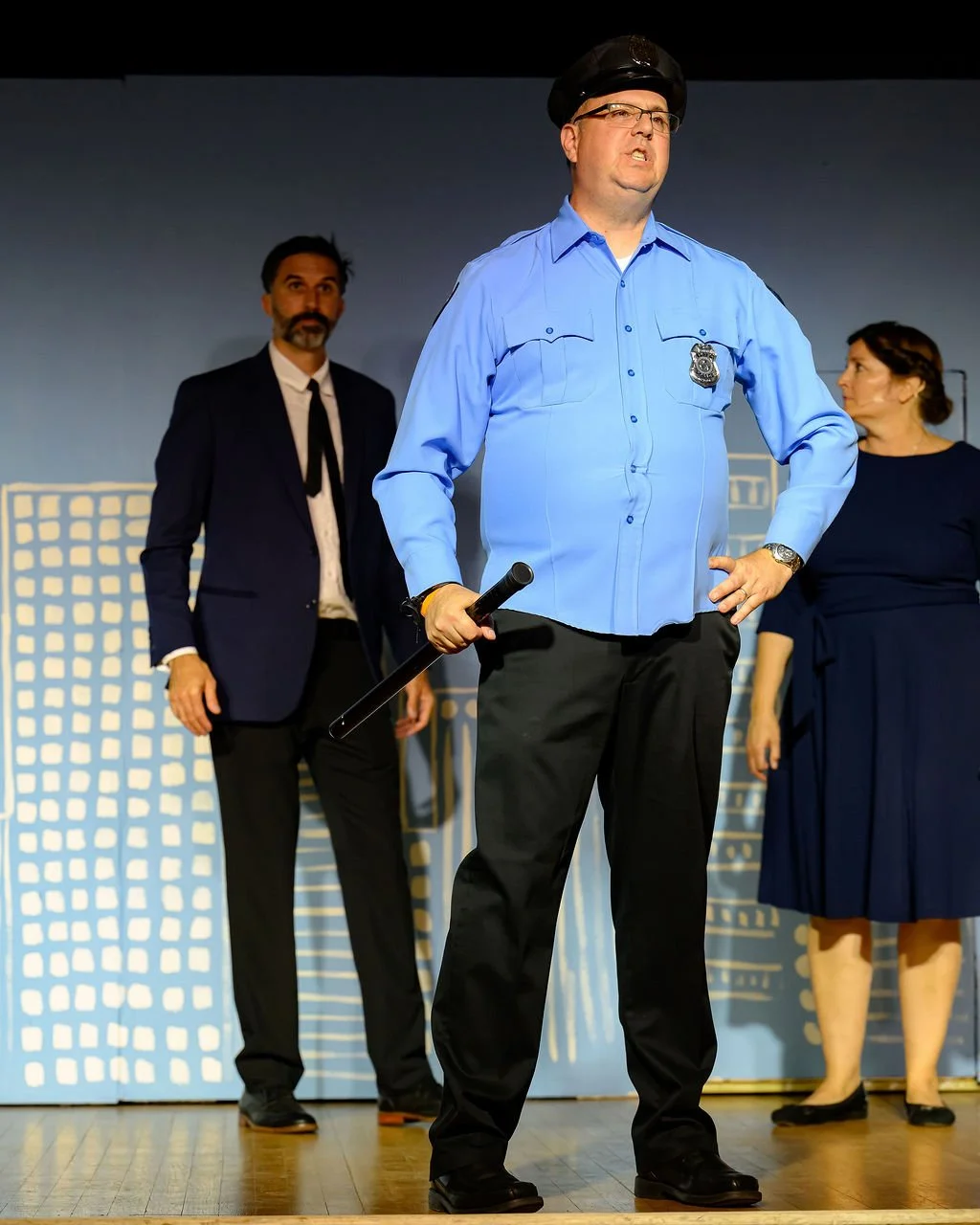 Man dressed as a police officer holding a baton, with two people in business attire standing behind him on a stage. The background features a cityscape drawing.