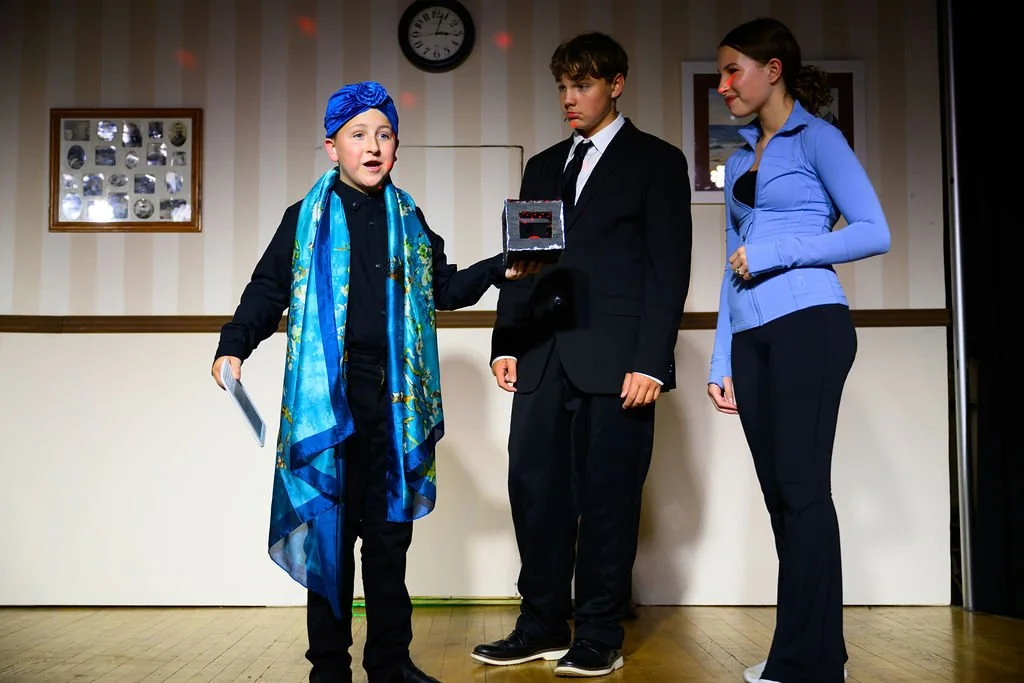 Three young actors on a stage performing a play, with one in costume holding a box, the other two in formal attire, in front of a beige wall with framed pictures and a clock.
