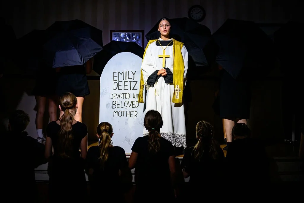 A young woman dressed as a nun or religious figure standing on stage in front of a crowd, holding a gold cross. Behind her are large black umbrellas and a gravestone prop with the name "Emily Deetz" and the words "Devoted Wife Beloved Mother" written