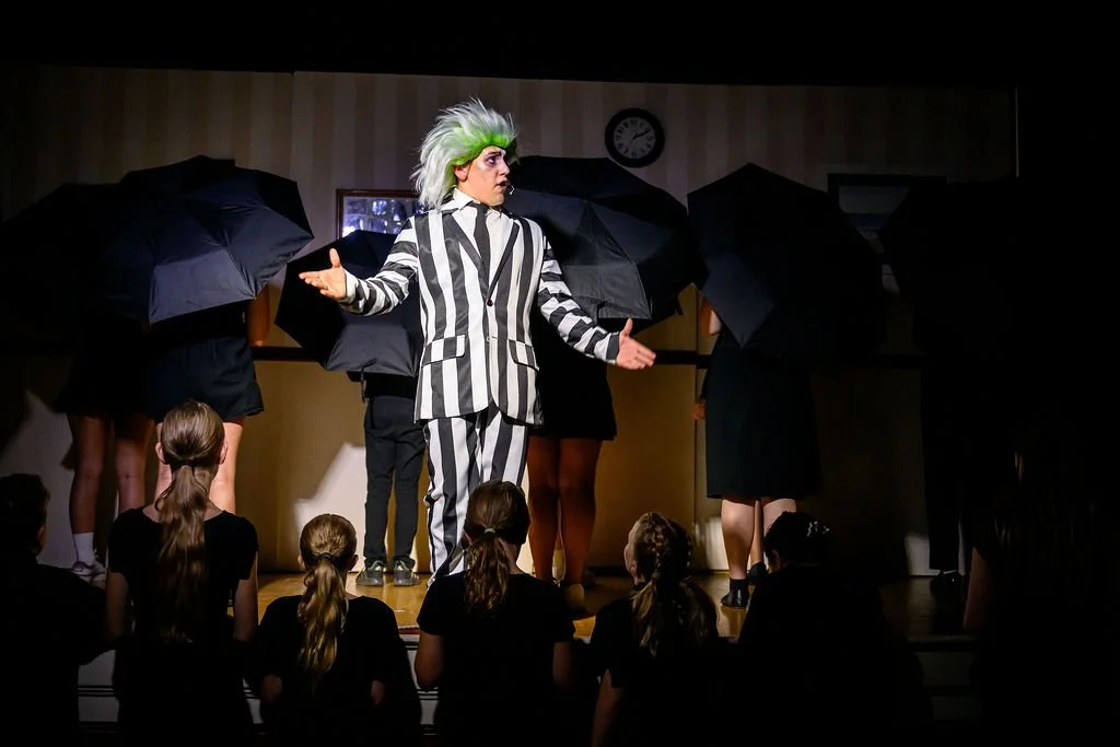 A theatrical performance featuring a person with a wild green and white wig dressed in a black and white striped suit, standing center stage with their arms open. Several people behind and around them hold black umbrellas, and the audience of childre