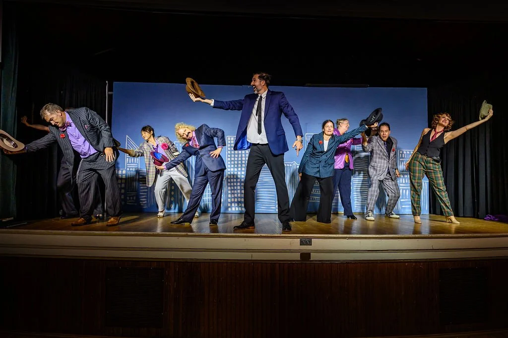 A group of performers on stage dressed in colorful business attire, holding hats, and striking dynamic poses during a theatrical performance.