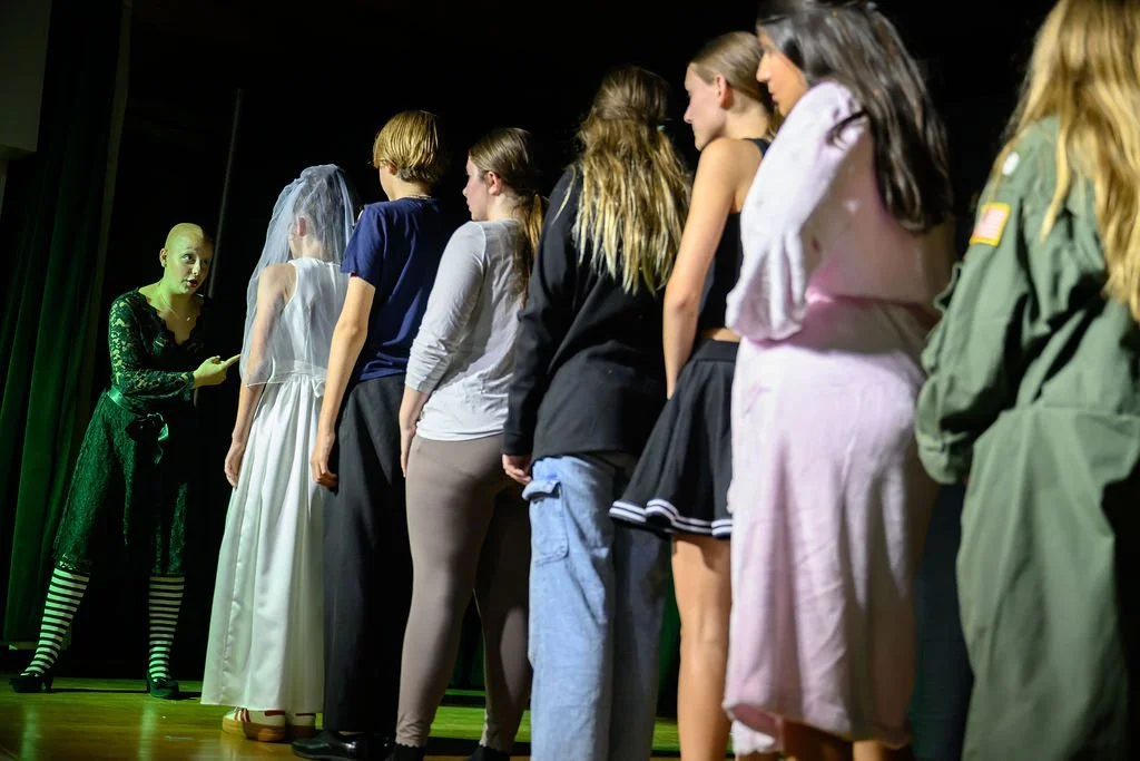 A woman in a black lace dress standing on stage in front of a line of children, some in casual clothes, some in costumes, during a theatrical rehearsal or performance.