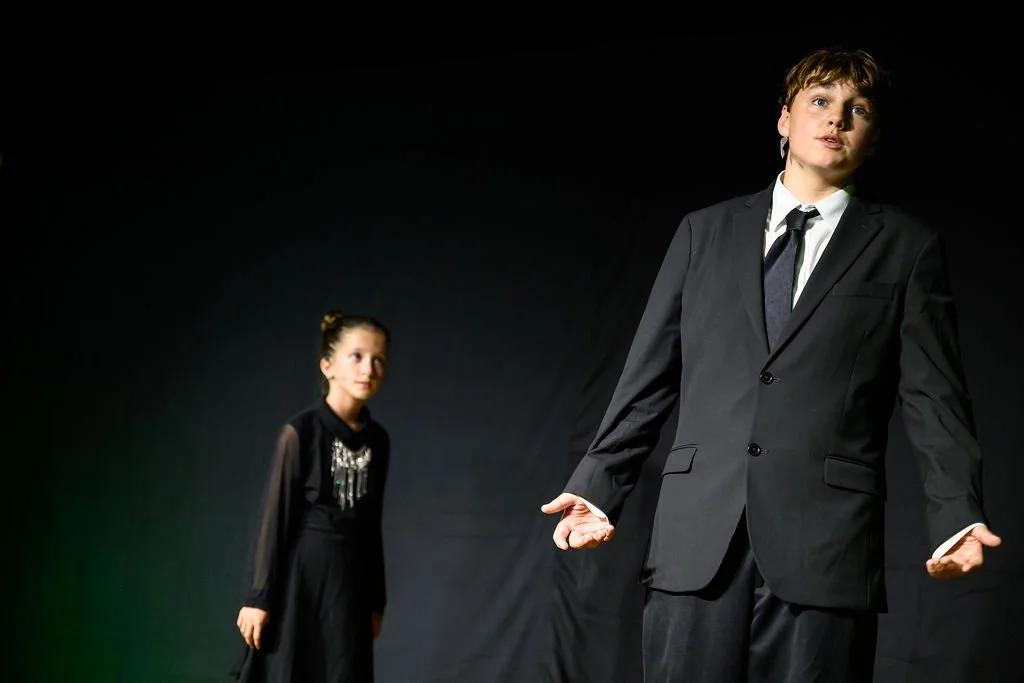 A young girl and a young boy on stage, with the boy in the foreground gesturing with both hands and wearing a black suit, while the girl stands in the background wearing a black dress with silver details, both against a dark backdrop.